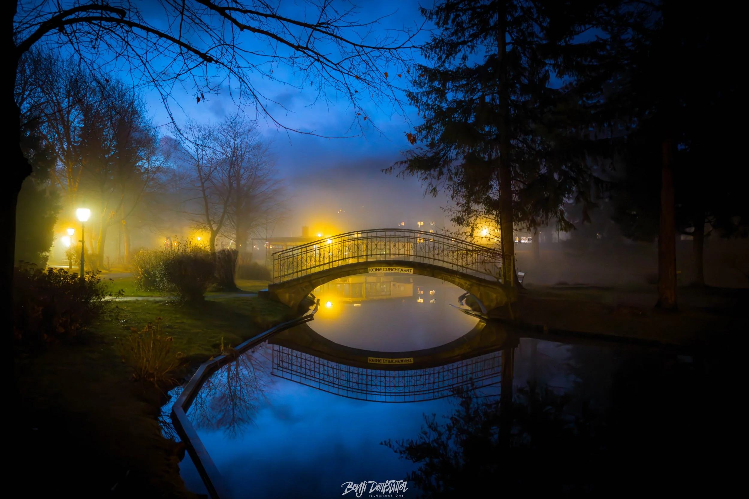 Nighttime scene of a park with a small arched bridge over a calm reflective canal, illuminated by street lamps and fog, with trees on both sides and a cloudy blue sky overhead.