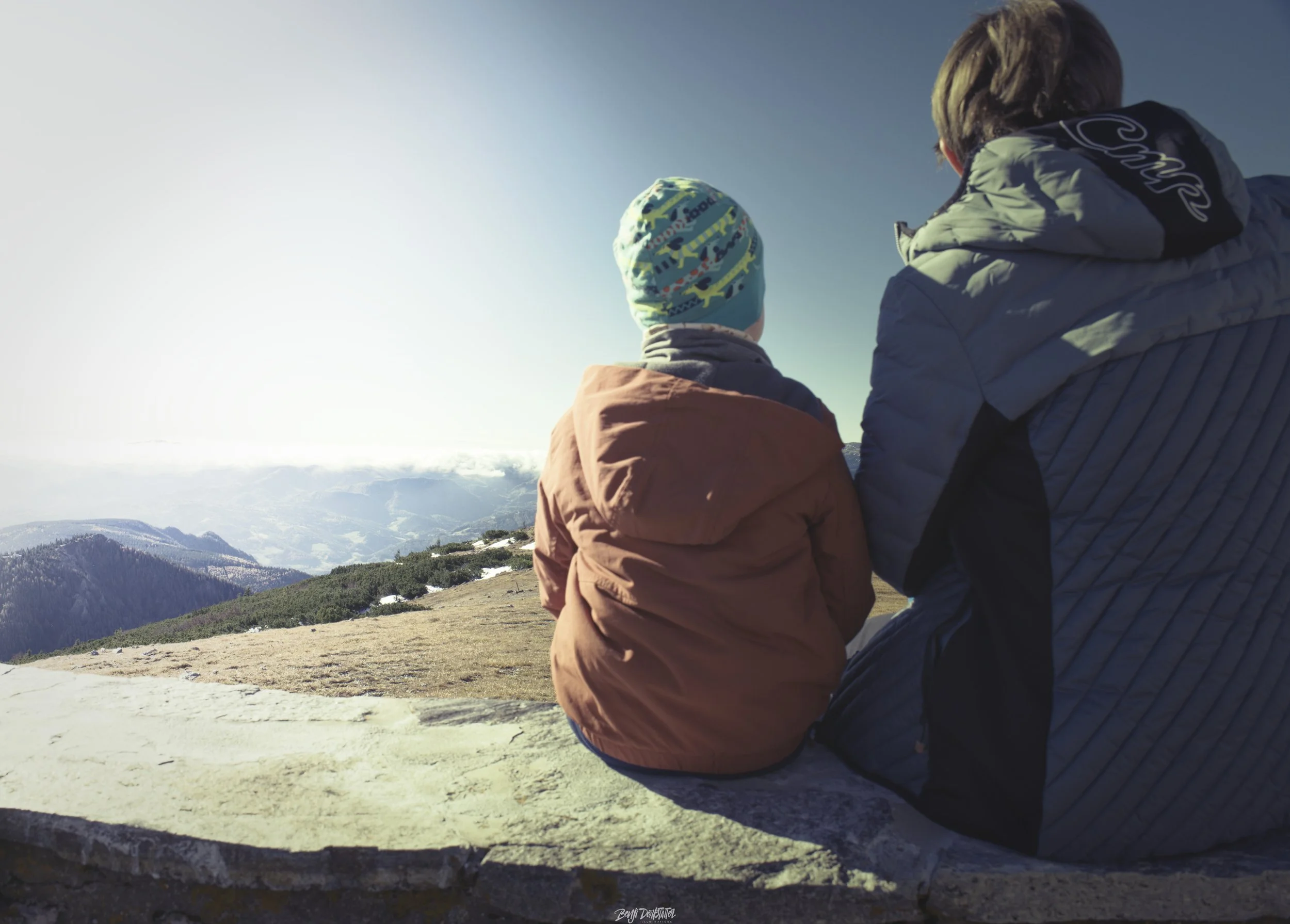 Two people, a child and an adult, sitting on a stone ledge looking at a mountain landscape during daytime. The child wears a colorful hat and orange jacket, and the adult is in a blue jacket with a hood.