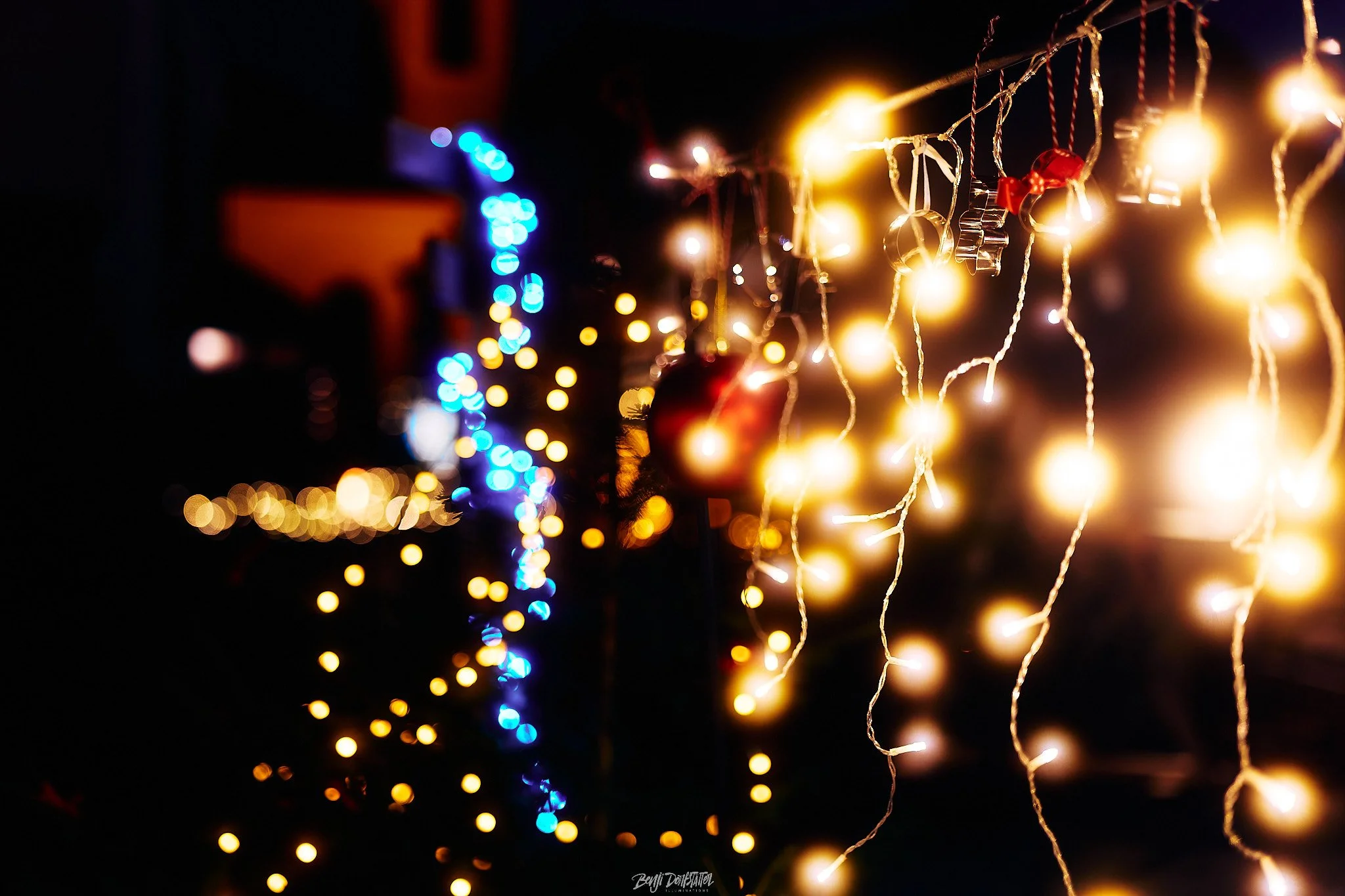 Close-up of outdoor Christmas decorations with warm yellow string lights and a red ornament, with blue and white lights blurred in the background at night.