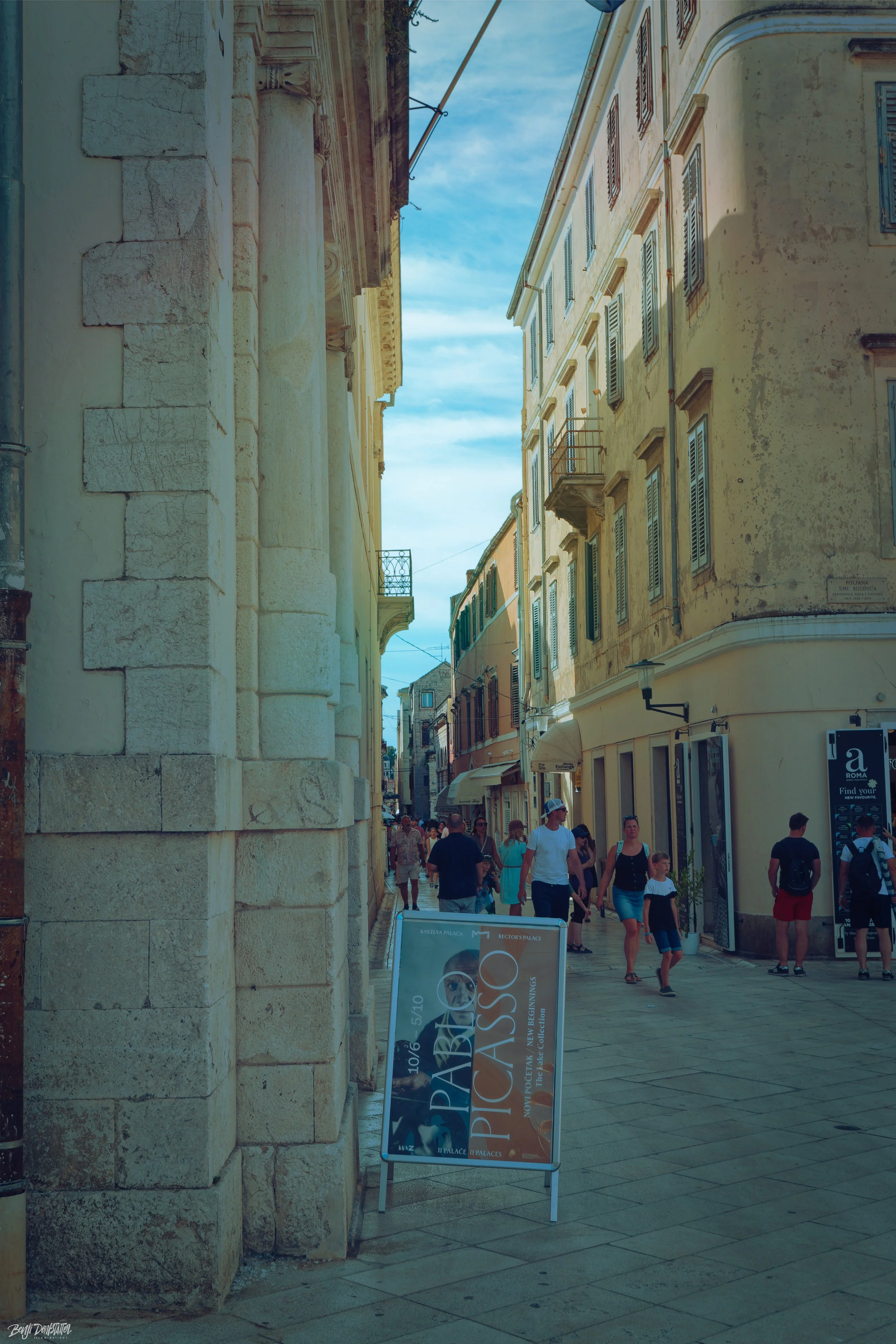 A bustling street in a historic European city with old buildings and a variety of shops. Pedestrians walk along the sidewalk with a sign advertising Picasso's exhibit in the foreground.