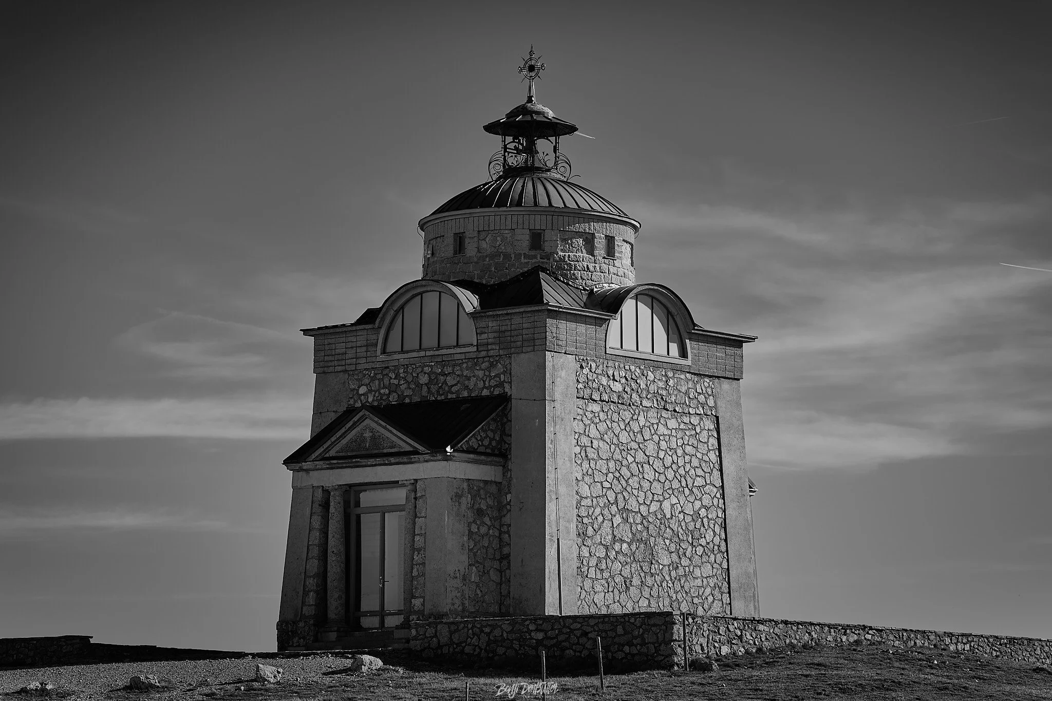 A black and white photo of a stone lighthouse with a domed roof and a weather vane on top, set against a cloudy sky.