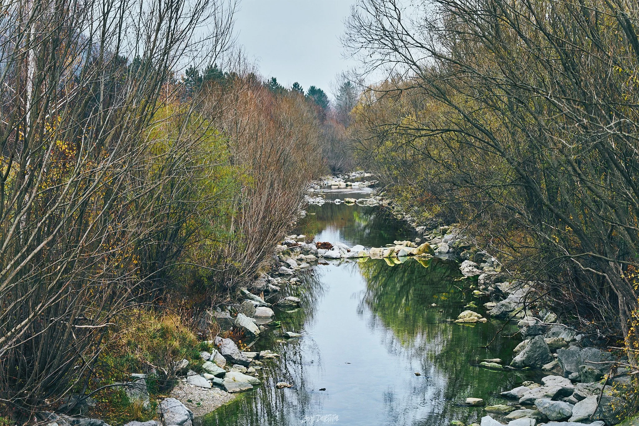 A narrow river lined with rocks and leafless trees and shrubs on both sides, with a cloudy overcast sky.