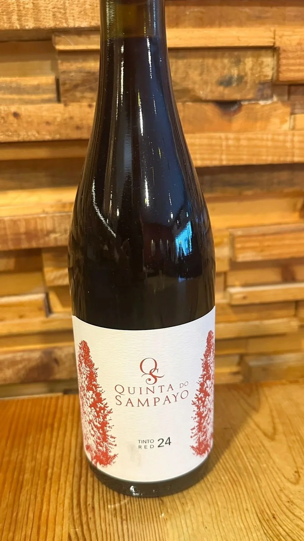 A bottle of Quinta do Sampeiro Colheita 2023 wine on a wooden surface with a wooden wall background.