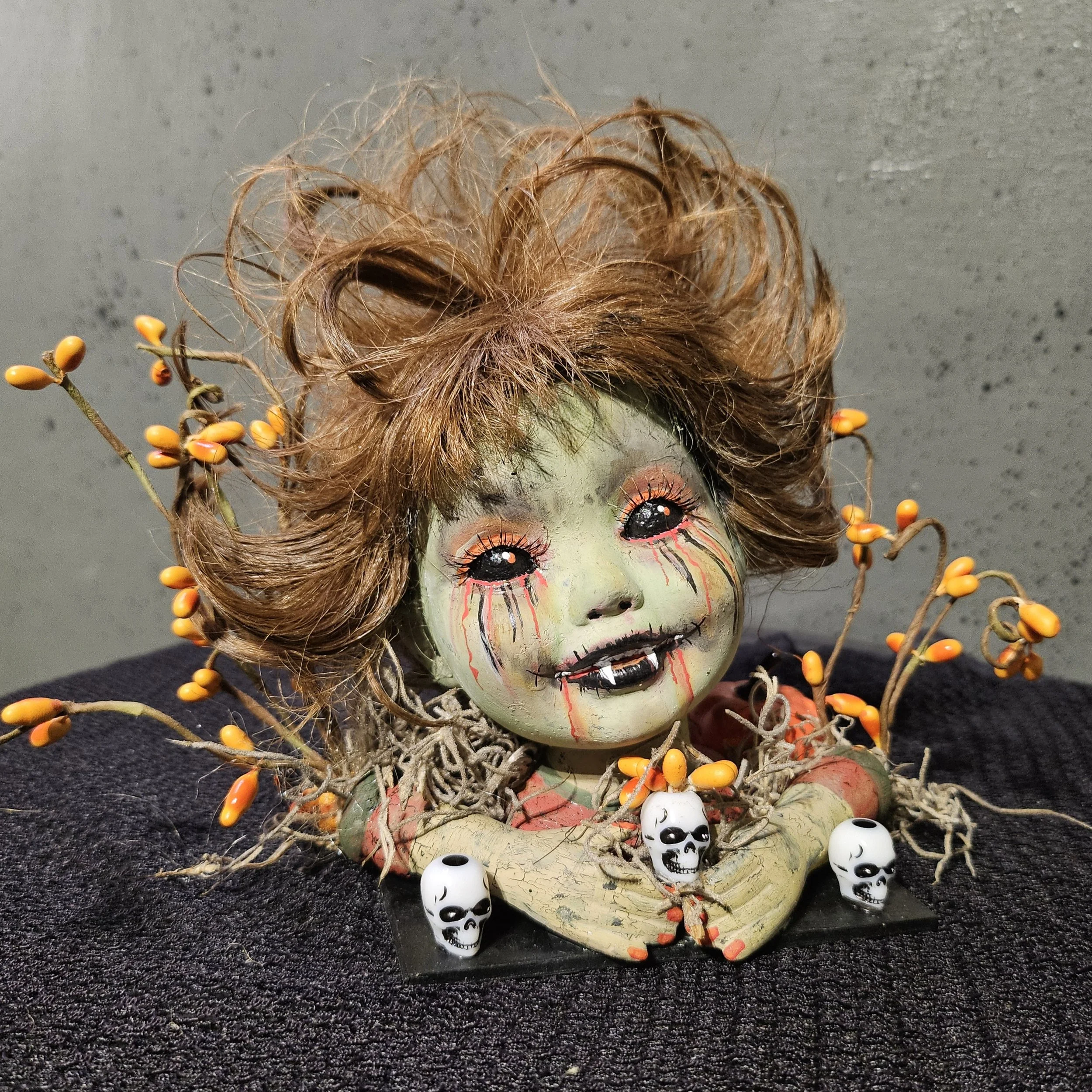  Front view of Autumn Bite, a gothic vampire doll surrounded by tiny skulls, pumpkins, moss, twisted branches, and orange autumn berries. 