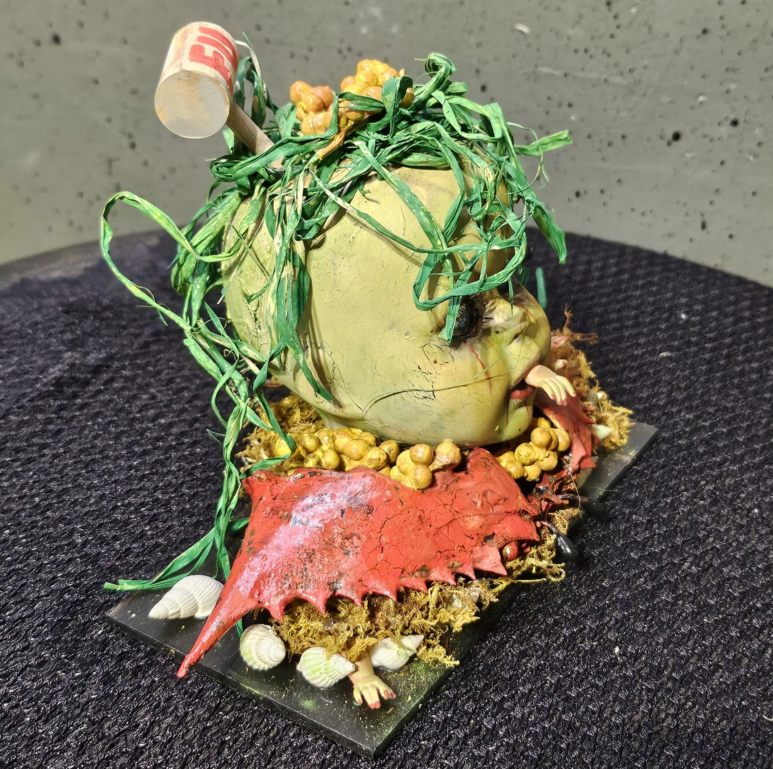  Right-side view of The Last Crab Boil showing the cracked doll head, tangled seaweed, shattered crab shell, miniature hands, seashells, and wooden mallet. 