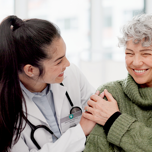 A female doctor smiling and holding an elderly woman's shoulder during a medical appointment in a bright clinic