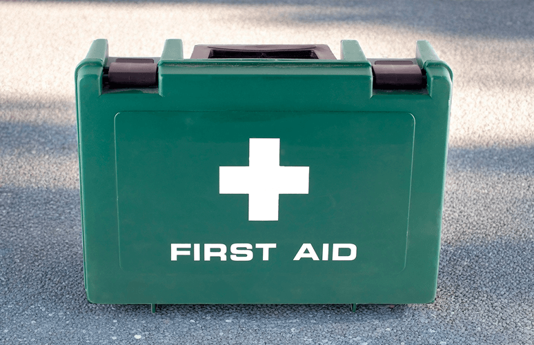 Green first aid kit with white cross symbol on the front, placed outdoors on asphalt.