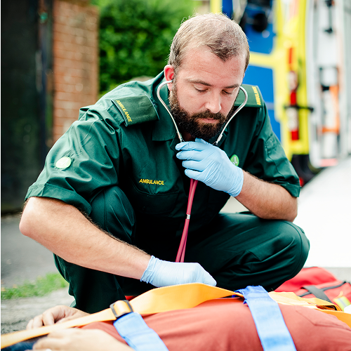 A male paramedic kneeling outdoors next to an emergency vehicle, inspecting a patient on a stretcher.