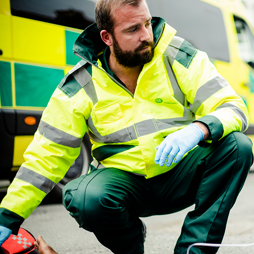 Emergency medical technician kneeling on the ground near an ambulance, wearing a yellow reflective jacket and blue gloves.