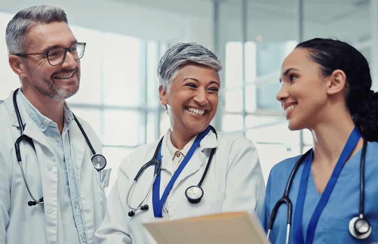 Three healthcare professionals, two women and one man, smiling and talking in a hospital. They are wearing medical scrubs and stethoscopes.