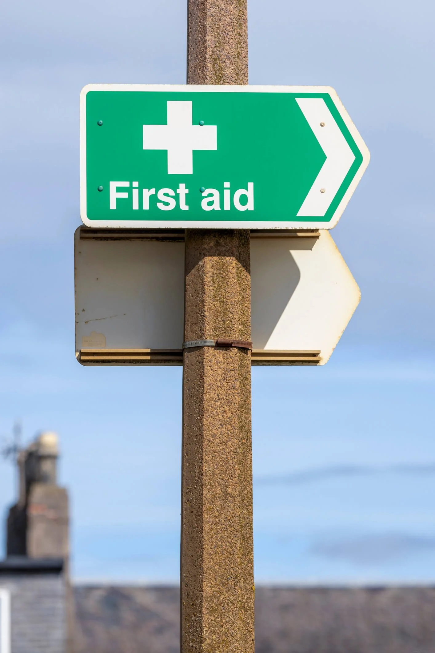 Green directional sign with a white cross and the words 'First aid' pointing to the right, mounted on a brown pole against a partly cloudy sky.