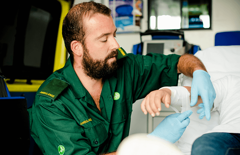 A paramedic in green uniform is tending to a patient, wrapping their hand with a bandage in a hospital or emergency room.