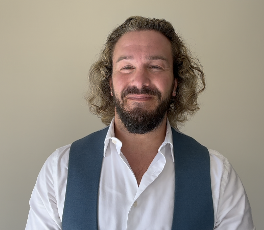 A man with curly hair and a beard smiling, wearing a white shirt and blue vest, standing against a plain light-colored background.