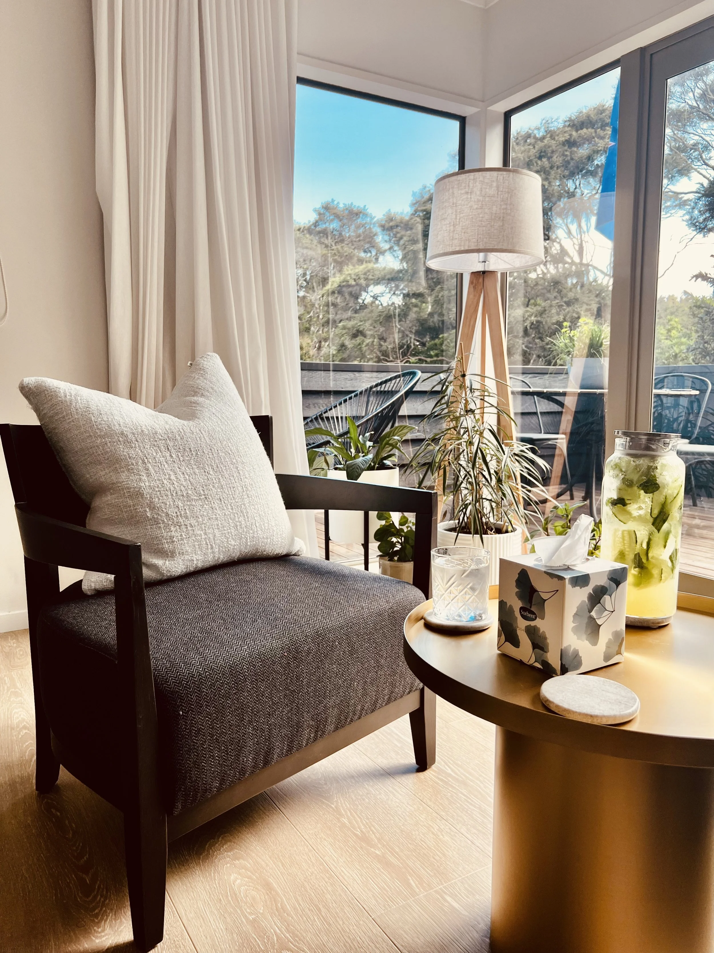 Cozy corner with black armchair, large white pillow, round wooden table with a glass of water, tissue box, container with infused water, surrounded by indoor plants, and a large window with white curtains letting in natural light.