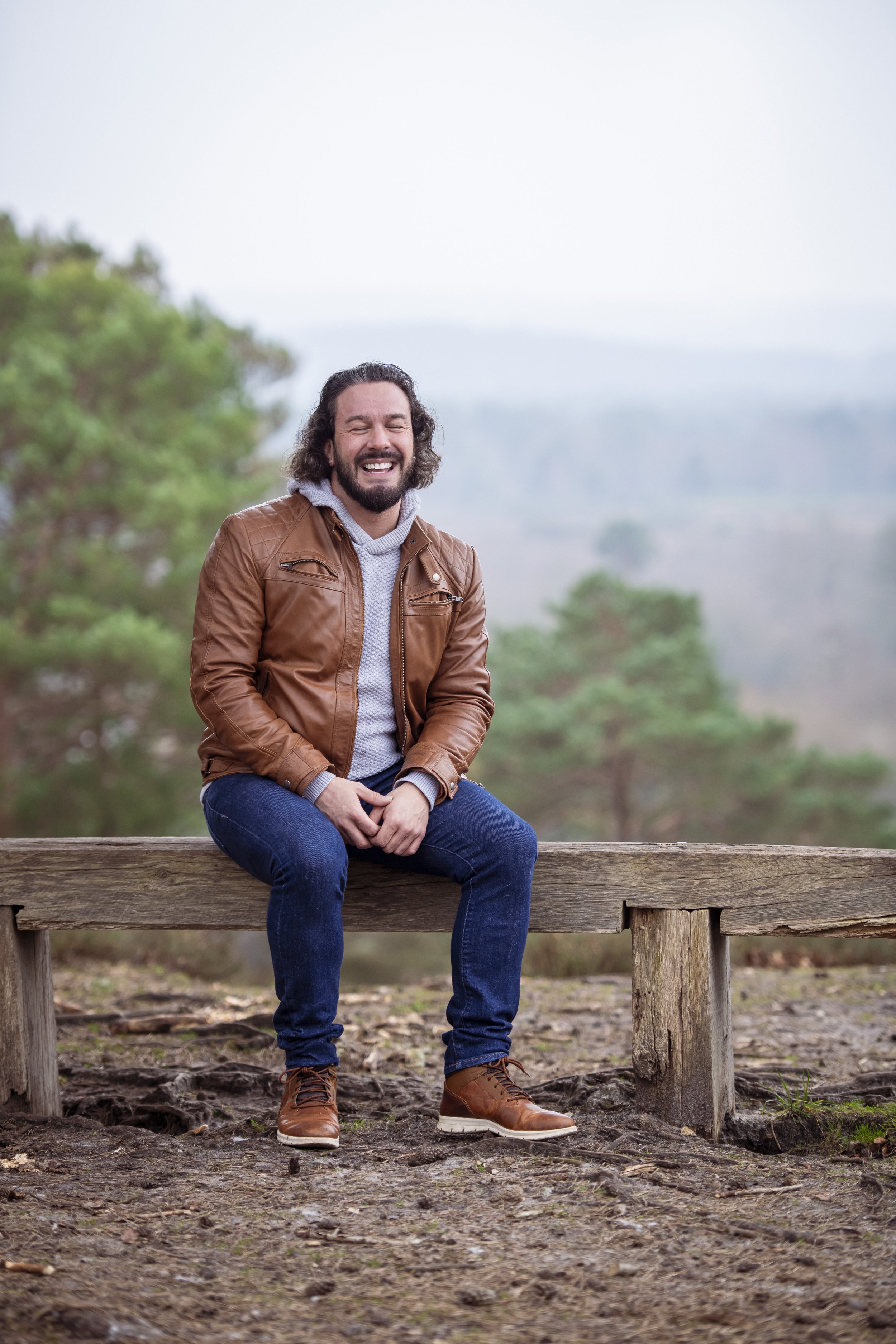 A man with long hair and a beard, wearing a brown leather jacket, gray hoodie, jeans, and brown boots, sitting on a wooden bench outdoors, smiling with his eyes closed.