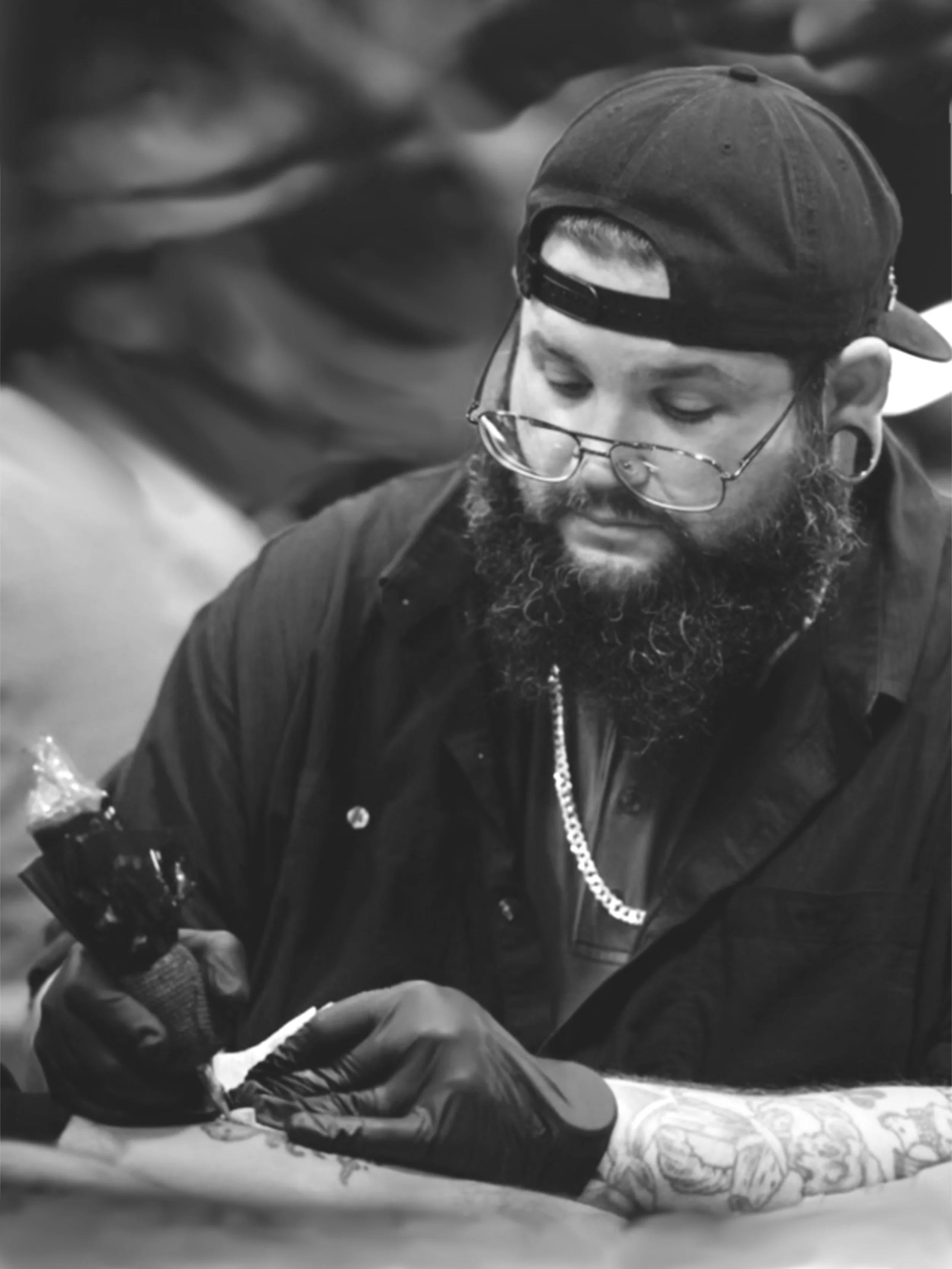 A man wearing a baseball cap backwards, glasses, and a chain necklace, with tattoos on his arm, tattooing. The man is focused on his work.