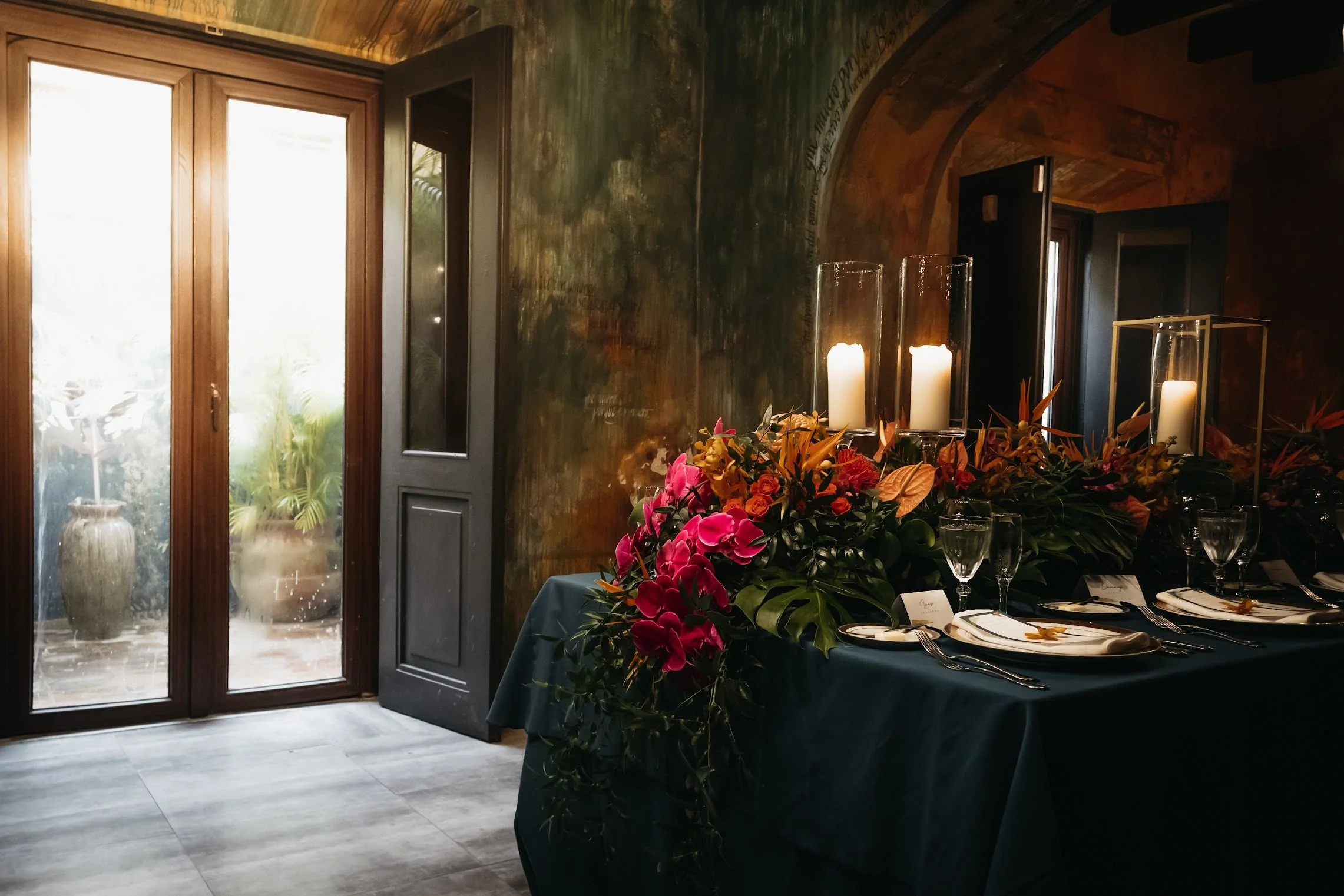 A dinner table decorated with pink and orange flowers, candles in glass holders, and place settings with plates, forks, knives, and glasses, in a dimly lit room with a wooden door and an open window.