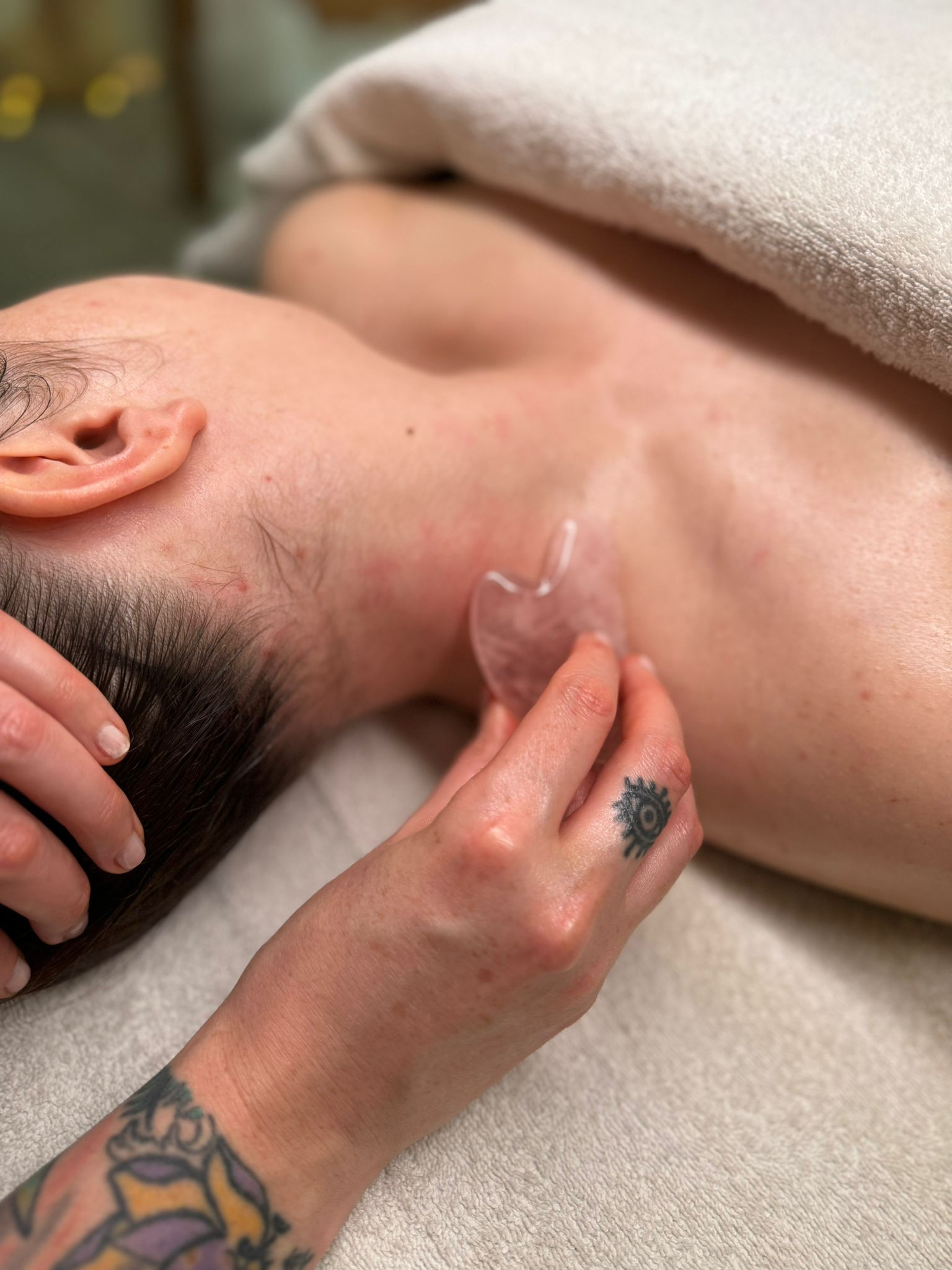 A person lying down on a massage table with a towel draped over their shoulder, receiving a gua sha facial massage using a clear stone tool on their neck. The person has dark hair, and the hand performing the massage has tattoos.