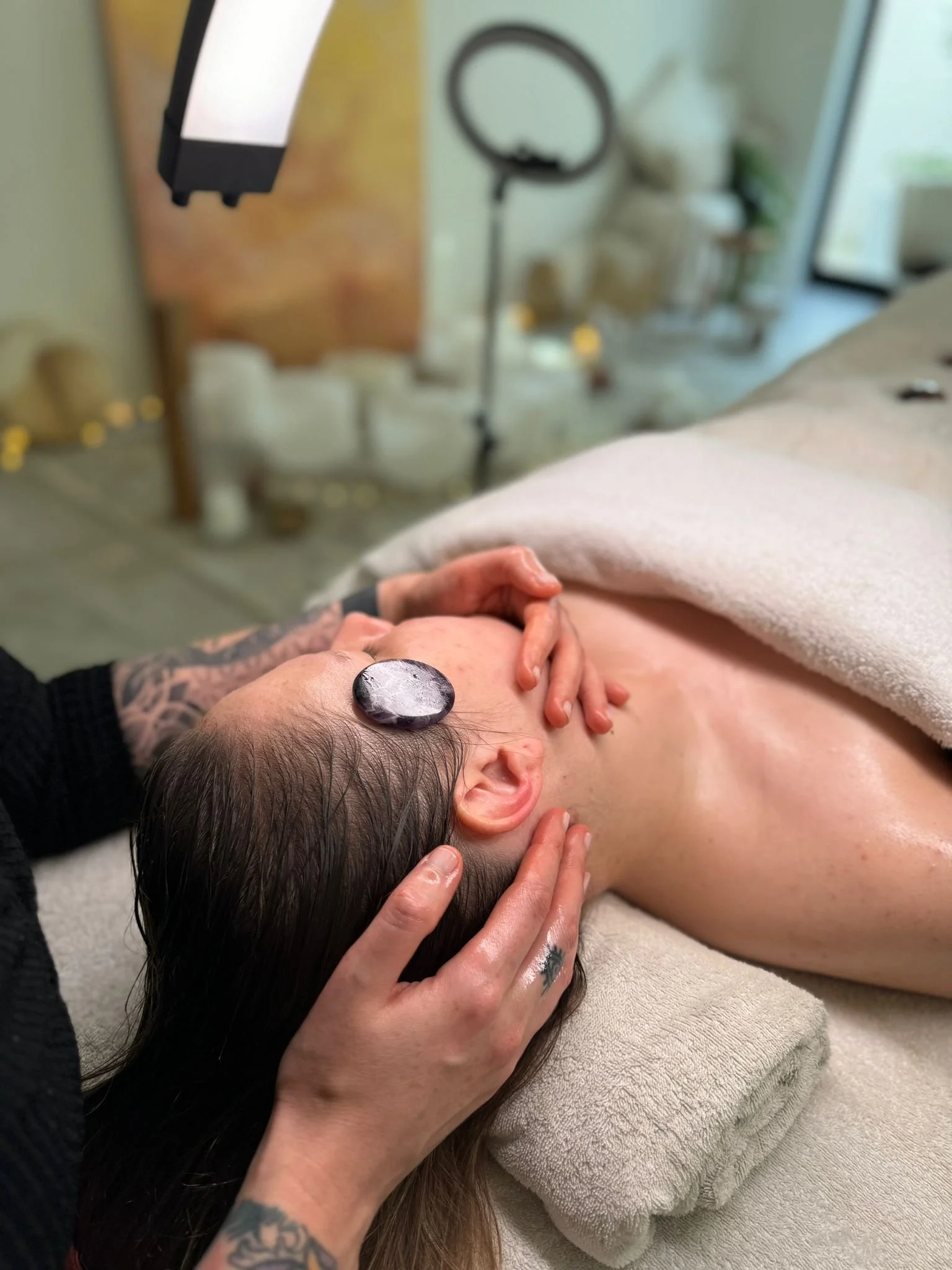 A person receiving a massage on their neck and shoulder area, with a massage stone placed on their head. The setting appears to be a spa or massage therapy room with towels, a ring light, and decorative items in the background.