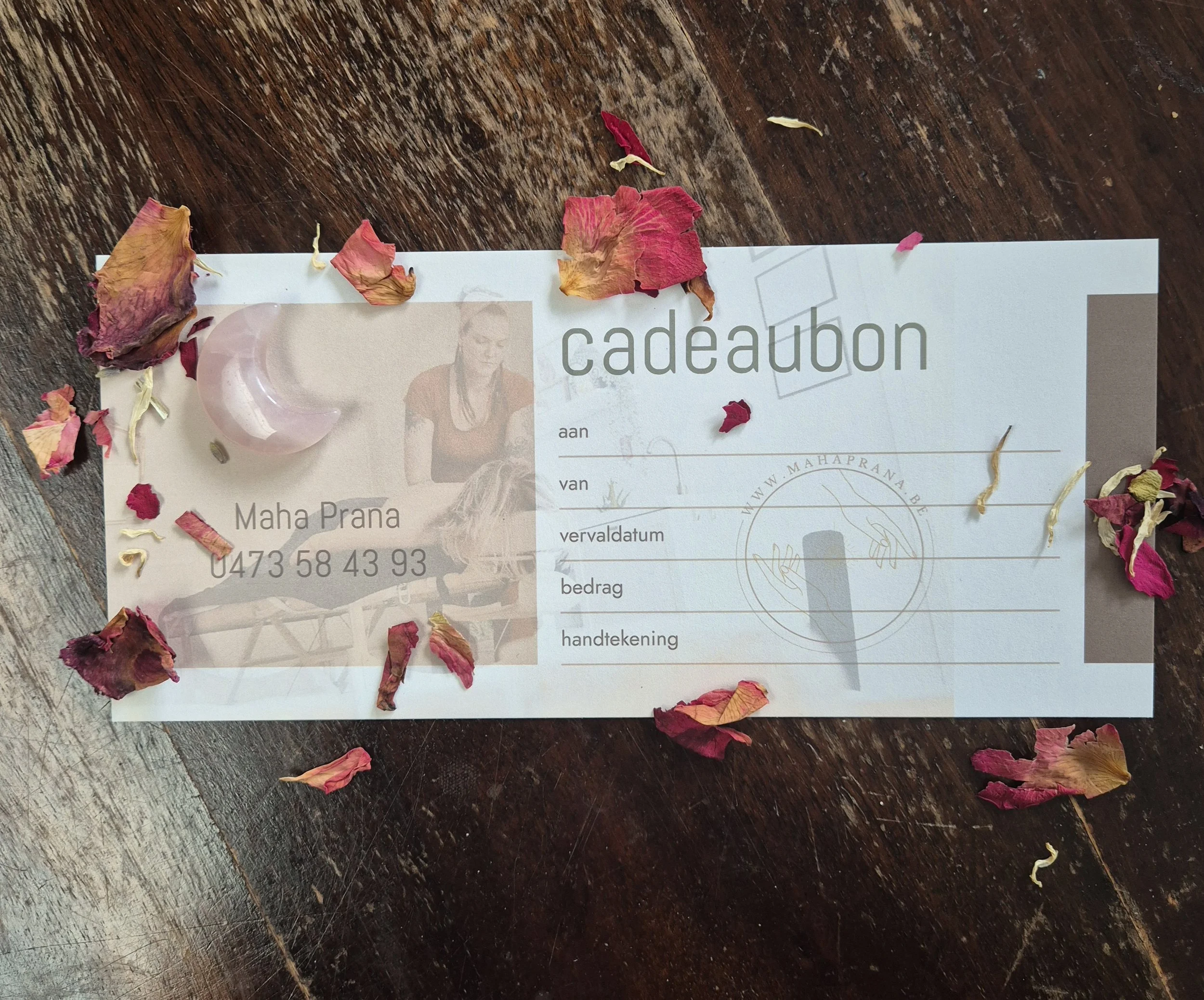 Gift voucher or gift certificate on a wooden surface, surrounded by scattered dried rose petals, with a crystal moon decoration placed on top.