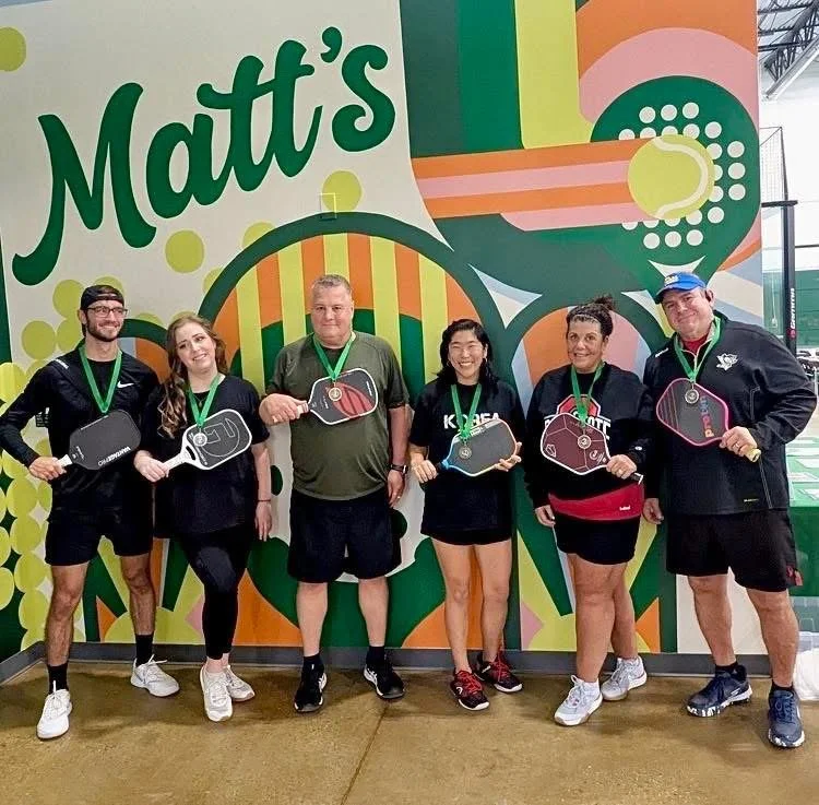 Pickleball players holding paddles in front of the Matt's Pickle and Padel mural with green medals after a tournament