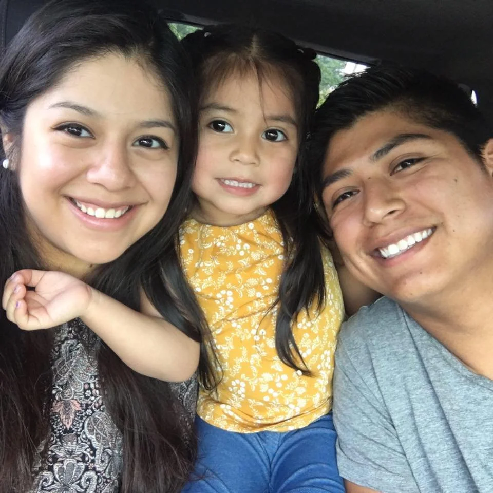 A smiling woman, a little girl in a yellow shirt, and smiling man posing closely for a selfie inside a vehicle.