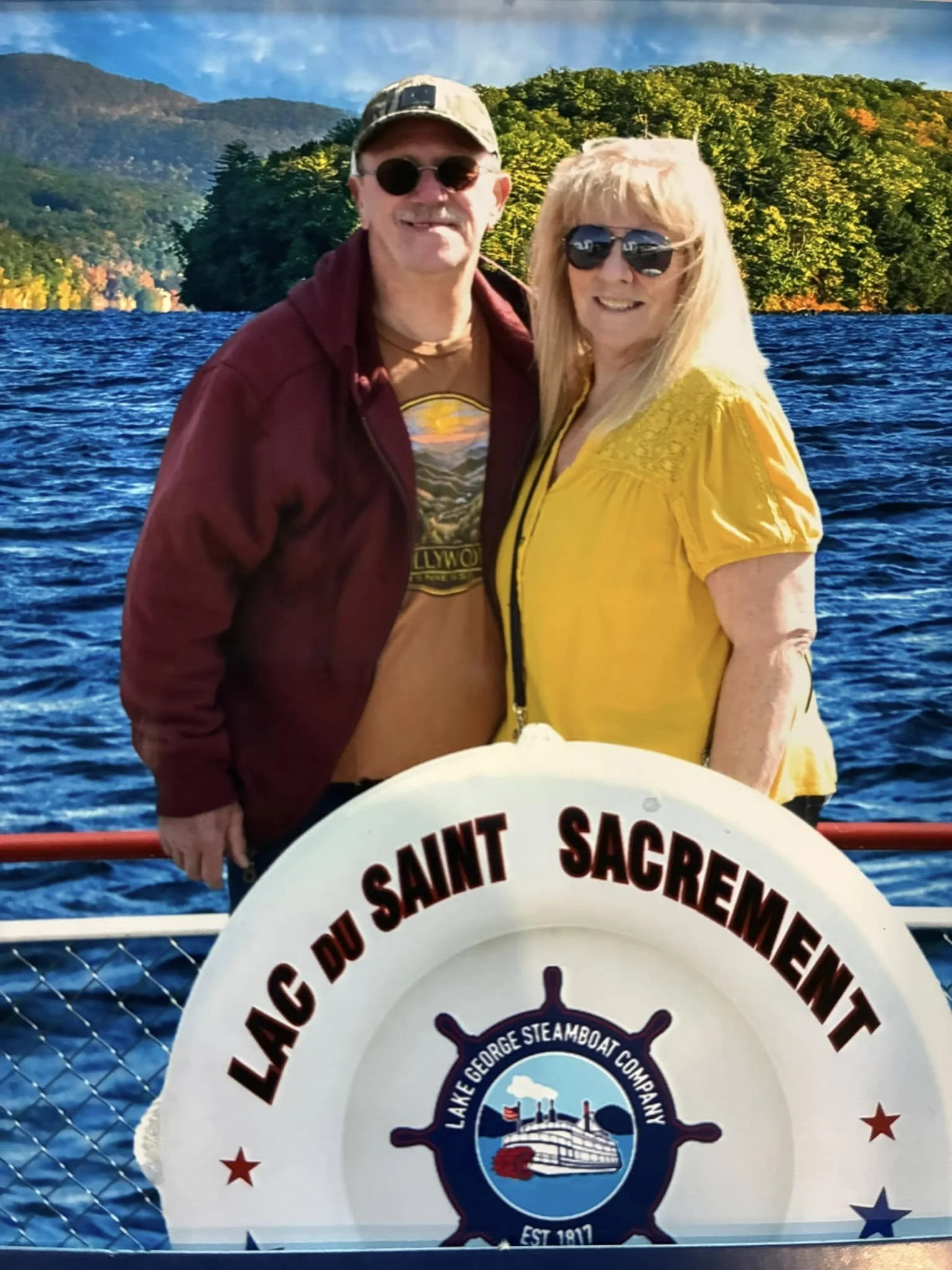 A man and woman smiling on a boat at Lake Saint Sacrement, with mountains and trees in the background. The boat features a life preserver with the logo of Lake George Steamboat Company.
