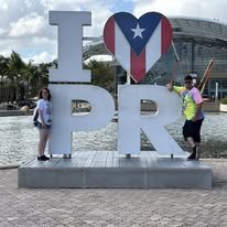 People posing next to a large 'I Love PR' sign with a heart shaped Puerto Rican flag in the center, outdoors with a domed building in the background.