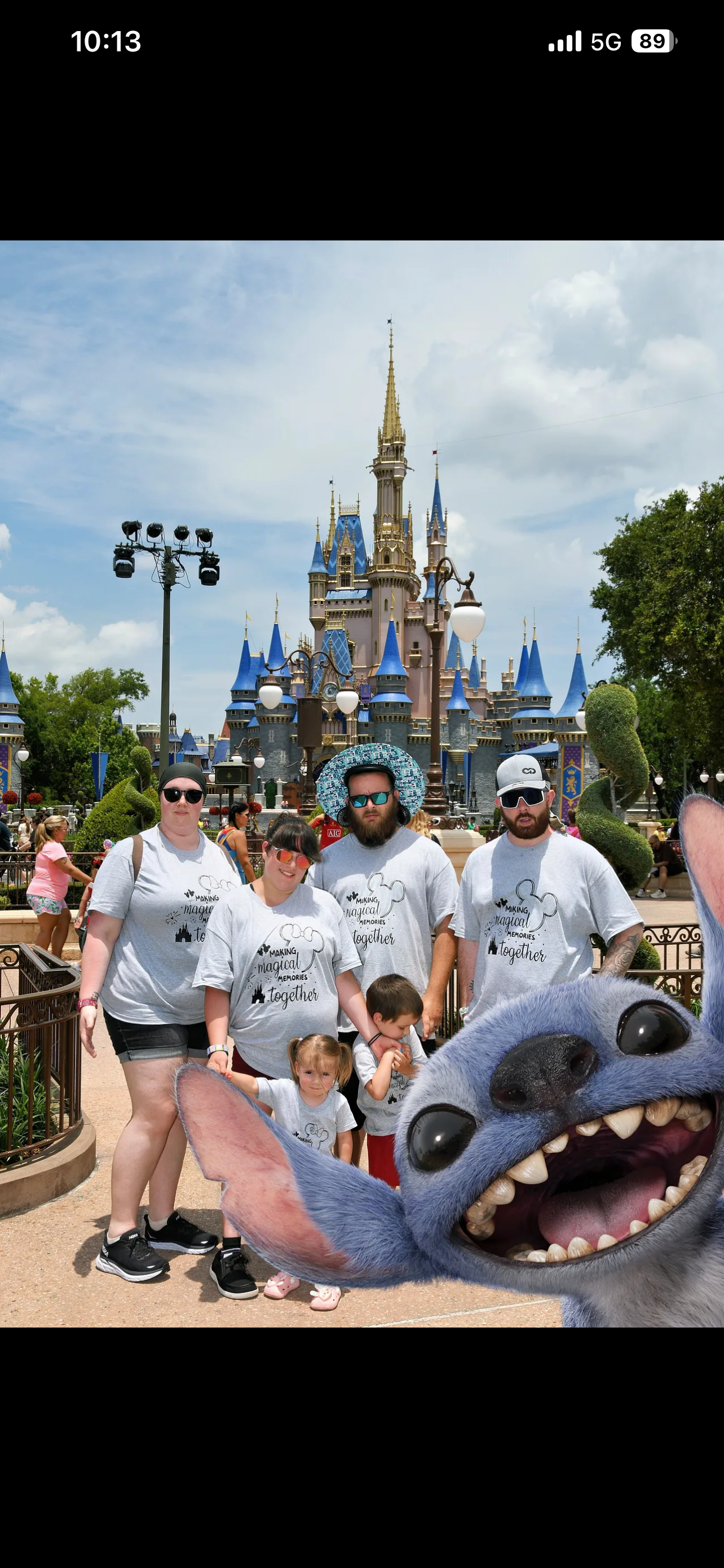 A family visiting Disneyland, with Sleeping Beauty Castle in the background. They are posing for a photo with a cartoon character, Stitch, in the foreground. The family is wearing matching T-shirts that read "Making Magical Memories Together." The setting is a sunny day with other visitors around.