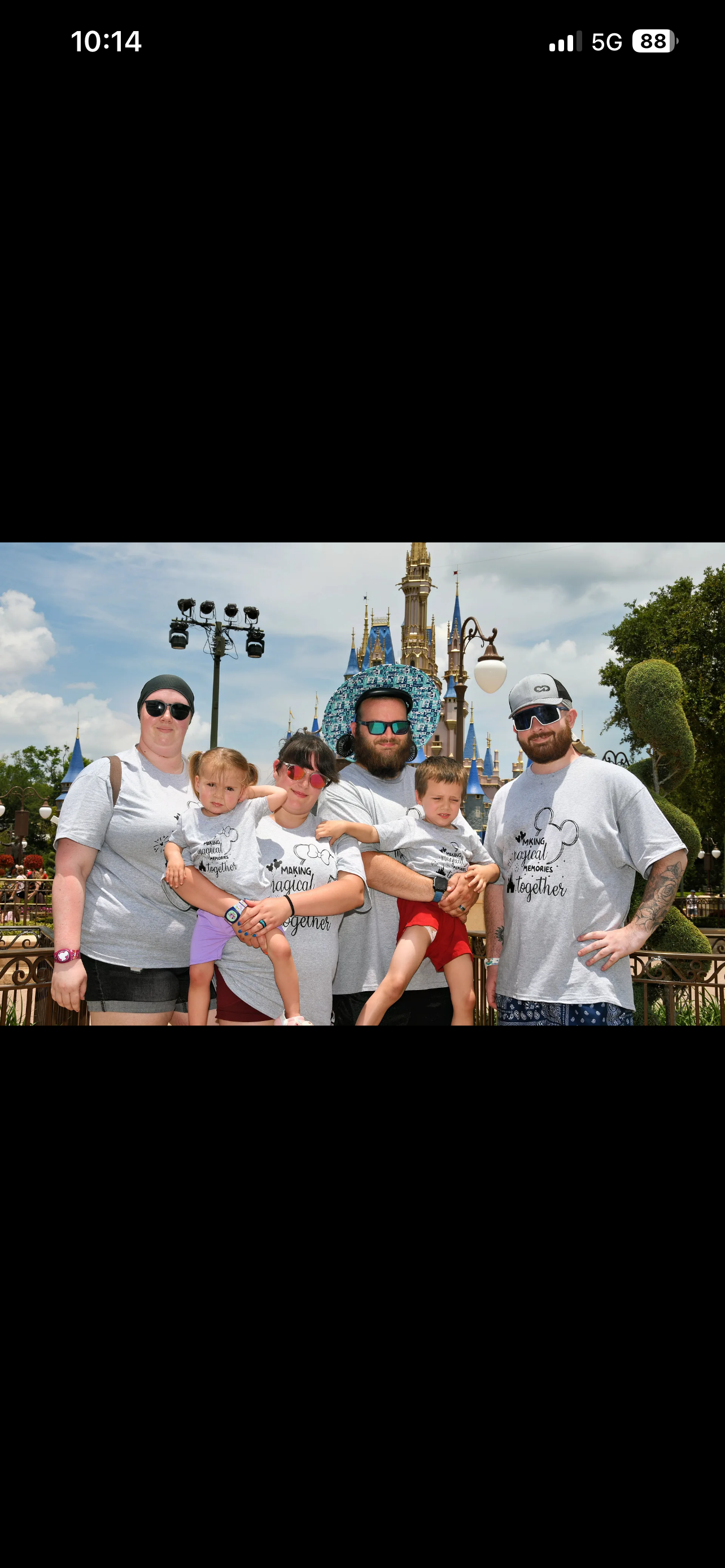 A family of five standing in front of Disney's Sleeping Beauty Castle at Disneyland, with trees and cloudy sky in the background, all dressed casually with matching Disney-themed t-shirts and sunglasses.