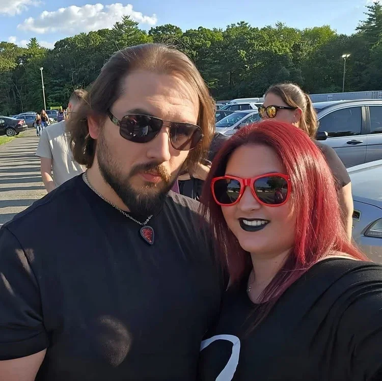 A man and woman taking a selfie outdoors in a parking lot. The man has shoulder-length brown hair, a beard, sunglasses, and is wearing a black shirt. The woman has long, red hair, red sunglasses, black lipstick, and a black shirt. Other people and cars are visible in the background.
