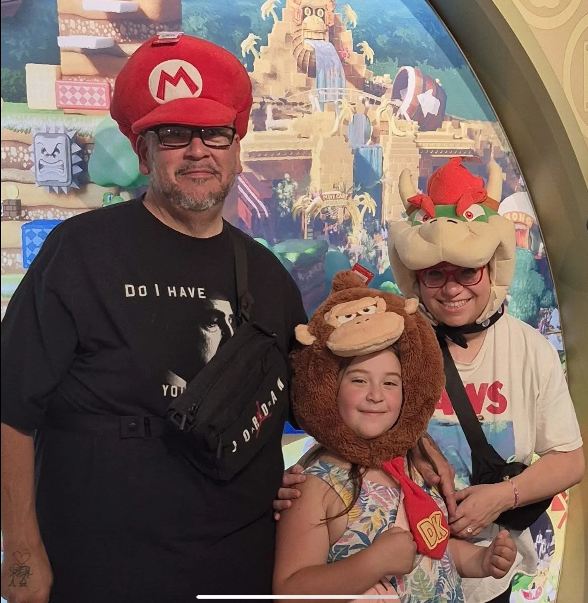 Three people in themed costumes from the Super Mario franchise posing together, with a colorful background featuring a fantasy landscape.