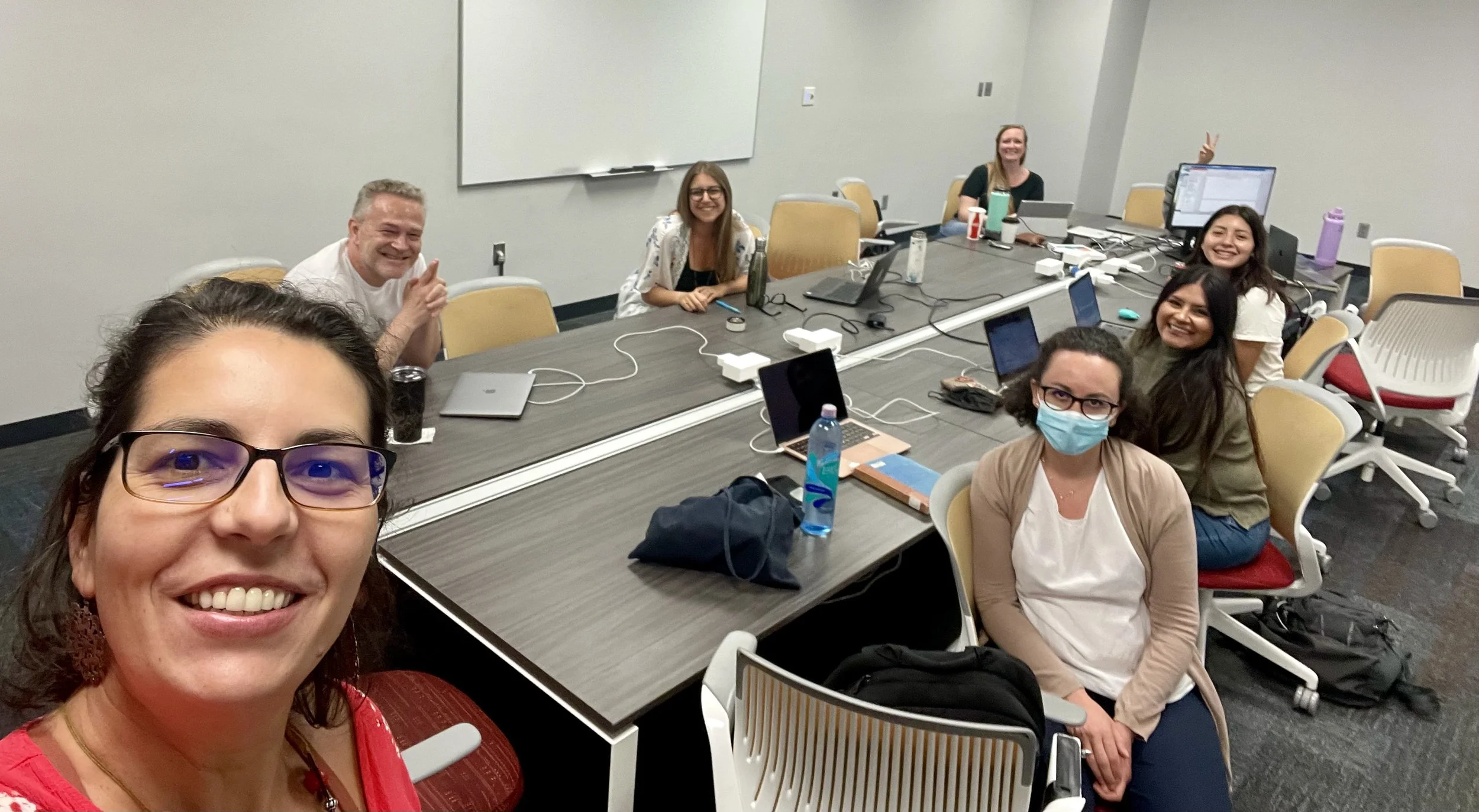 Group of diverse people in a conference room with Alyssa Birnbaum taking a selfie, some smiling, some wearing masks, with laptops and water bottles on the table.