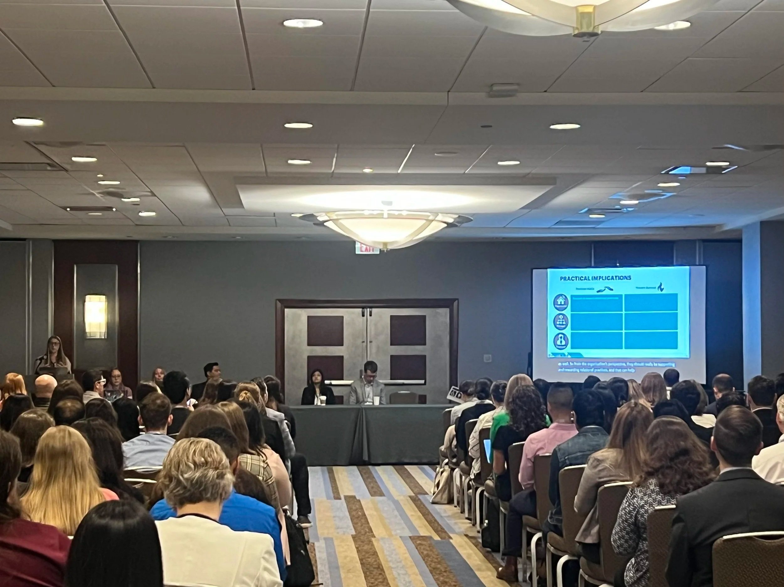 A large conference room filled with seated attendees watching a presentation on a large screen. The presentation slide titled "Practical Implications" features icons and text. There are panelists seated at a head table at the front of the room, and a woman standing on the left side near the wall.