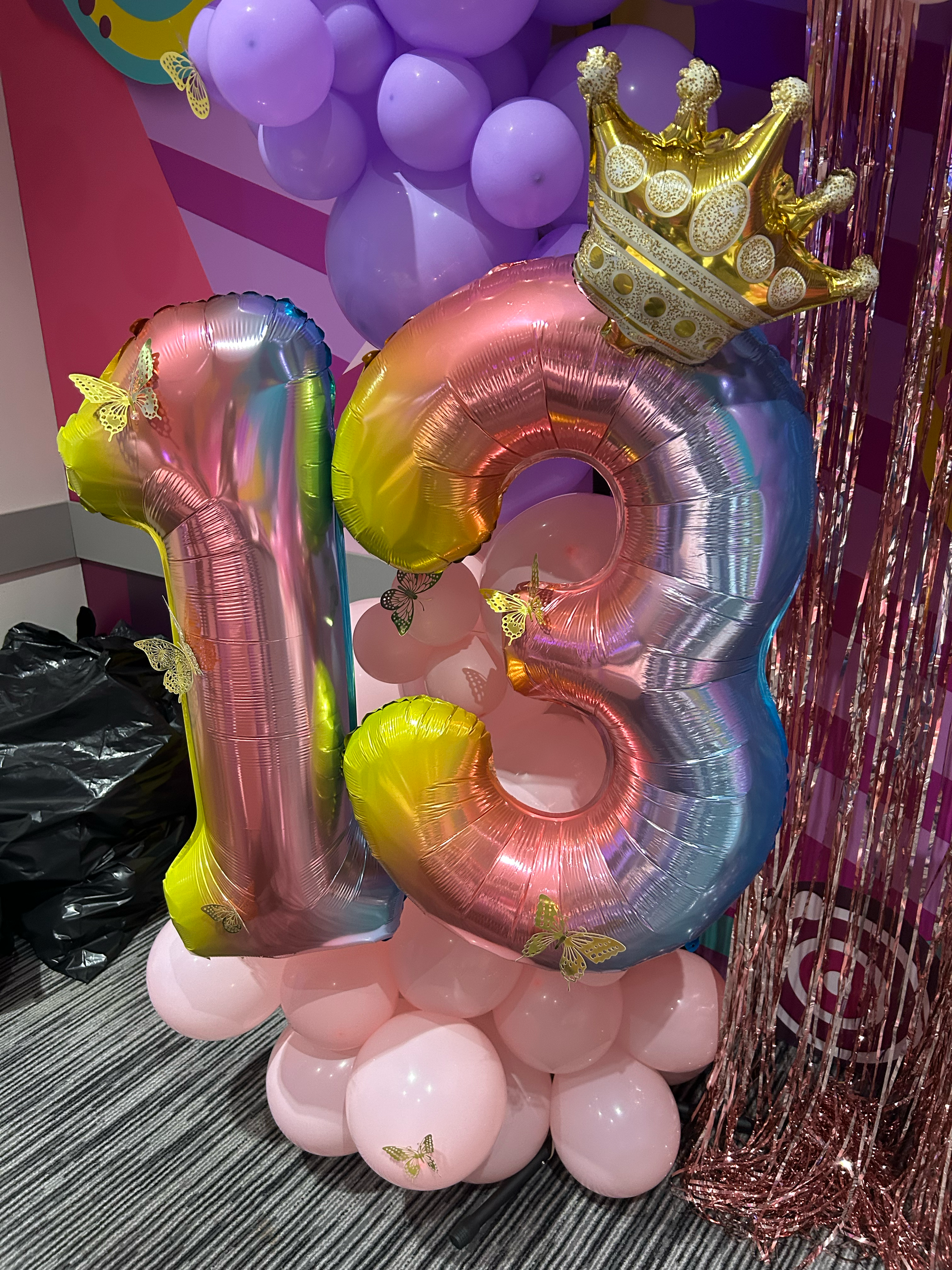 Number 13 birthday balloon arrangement with pink balloons at the base, large metallic number balloons in pink, yellow, and blue, and a gold crown balloon, decorated with yellow butterfly stickers, against a pink and purple themed backdrop.
