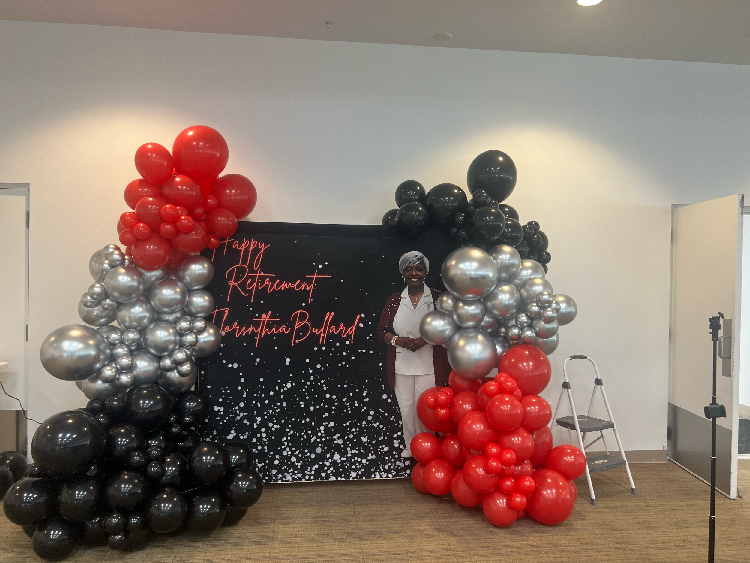 A woman in white pants and a white top, with a red jacket, standing in front of a decorated backdrop with balloons and a sign that says "Happy Retirement Lorinthia Bullard." The balloons are red, black, and silver, arranged around the backdrop.