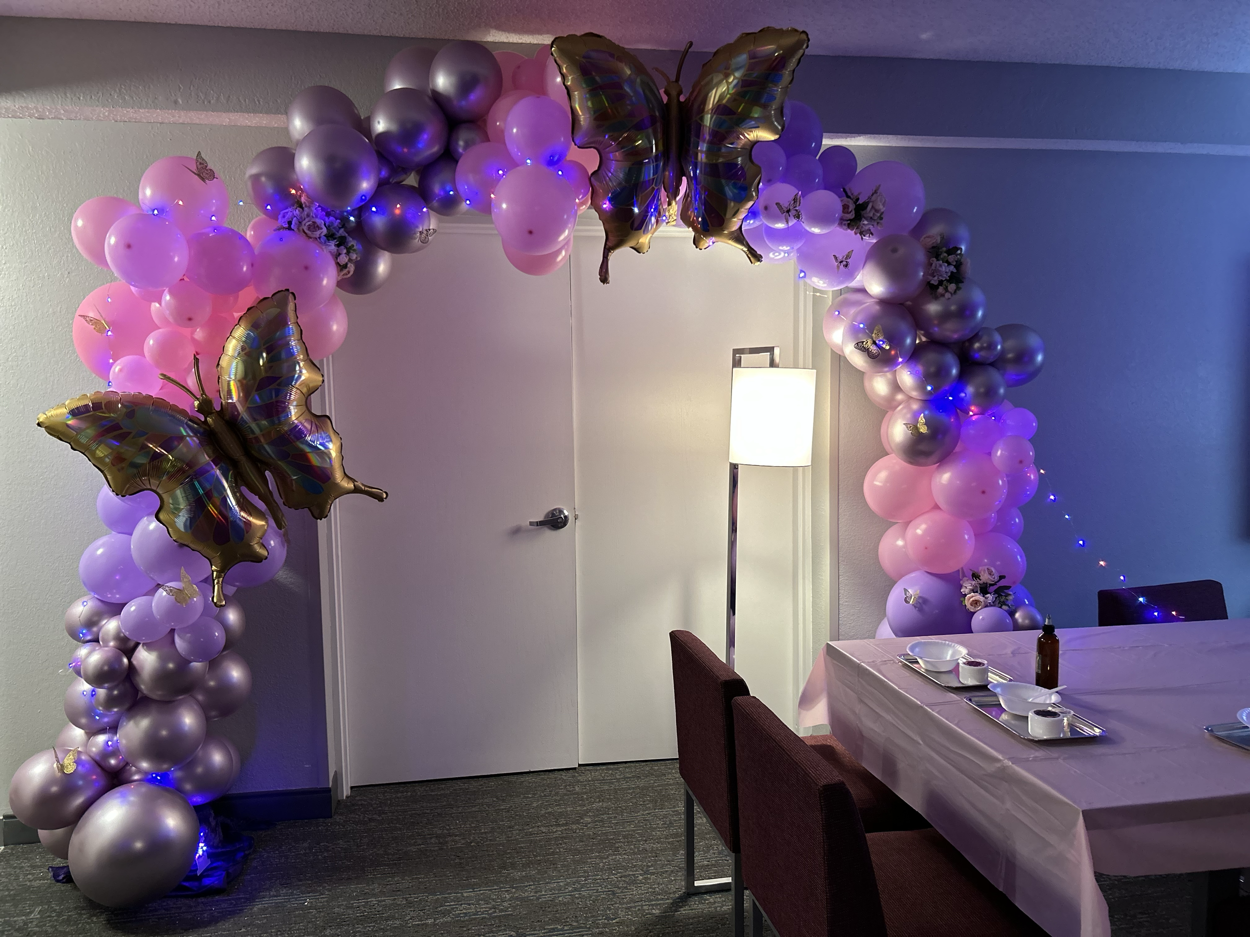 Balloon arch decorated with pink and purple balloons, butterfly-shaped balloons, and flower arrangements. It is set in a room with a table covered in a pink tablecloth and chairs, and a white door with a standing lamp nearby.