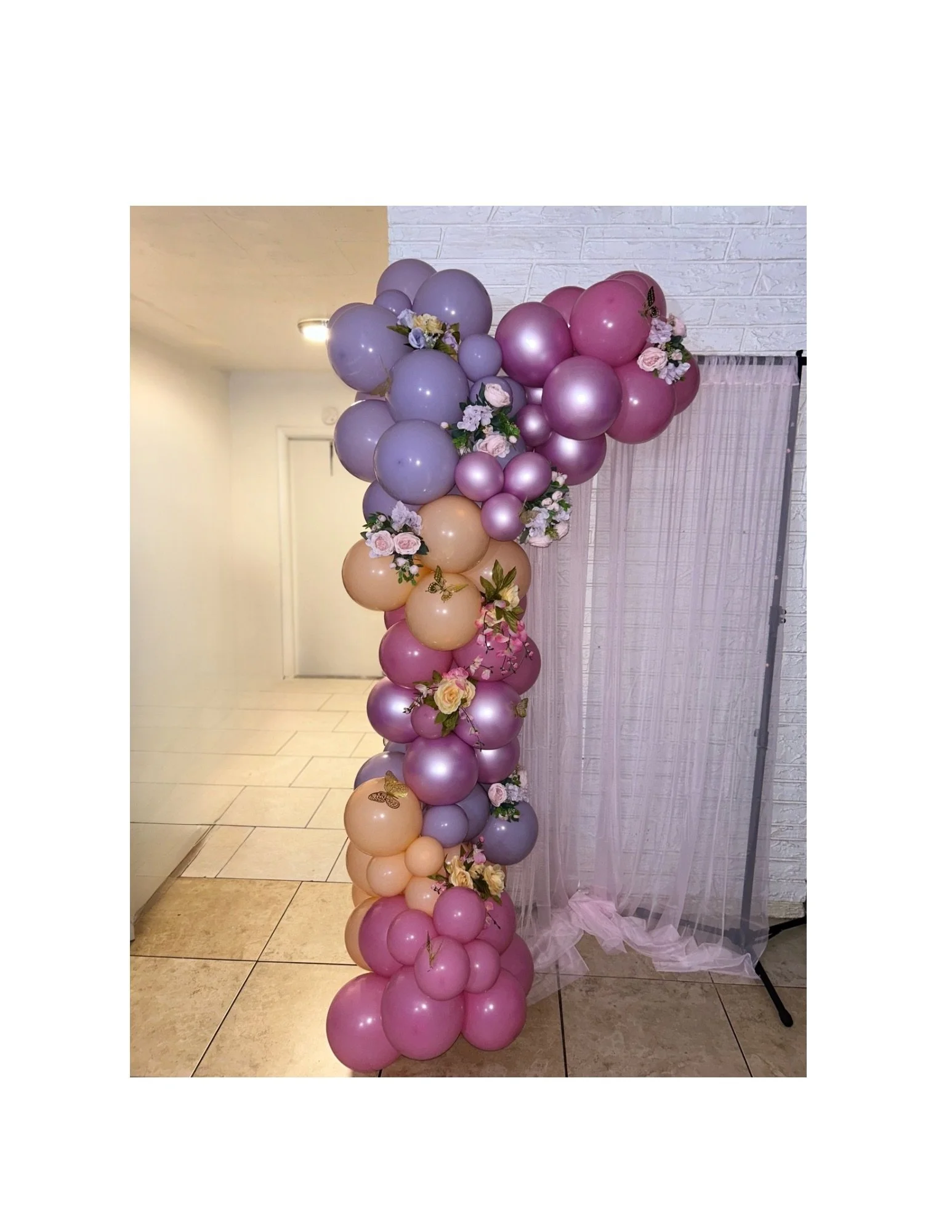 Balloon arrangement with pink, purple, and beige balloons decorated with white and pink flowers, set against a backdrop of sheer pink fabric and a white brick wall.