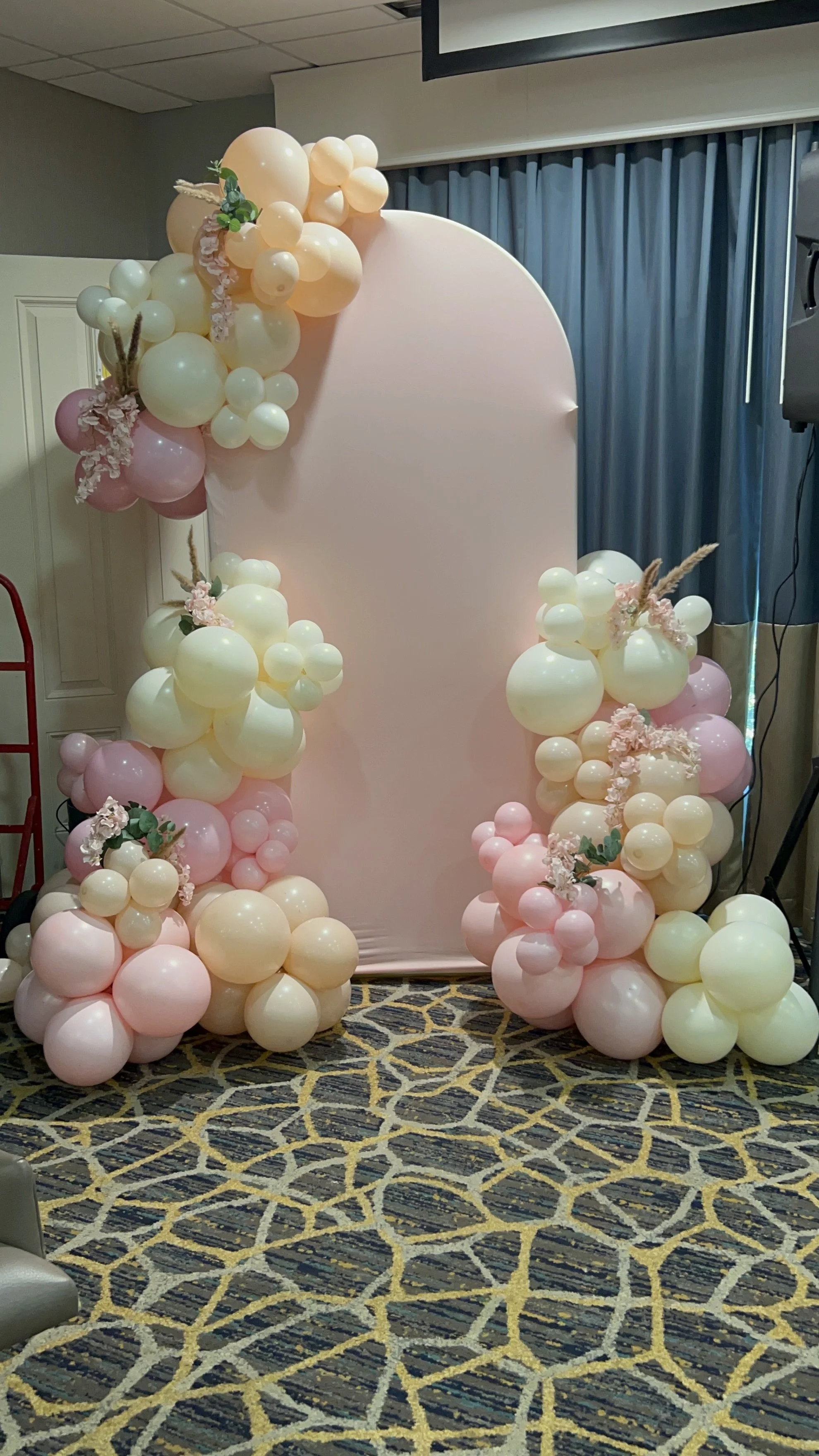 Decorative balloon arch with pink, cream, and white balloons, accented with pink flowers and beige pampas grass, arranged around a soft pink backdrop.