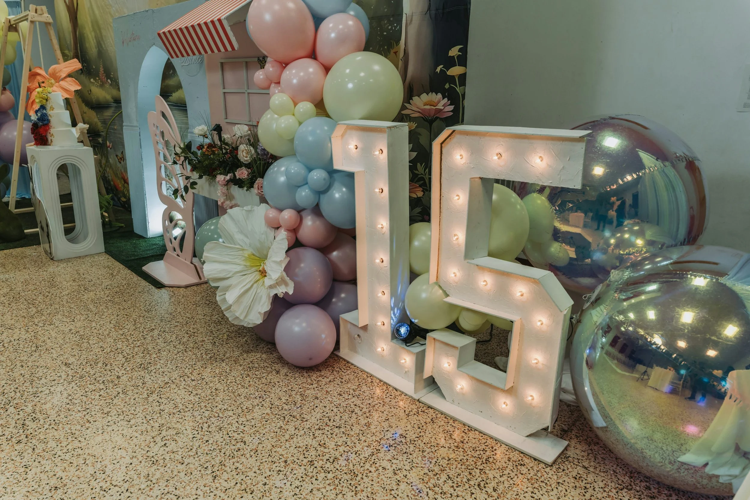 Decorative display for a celebration with pastel balloons, a large illuminated number '15', metallic balloons, and floral arrangements.
