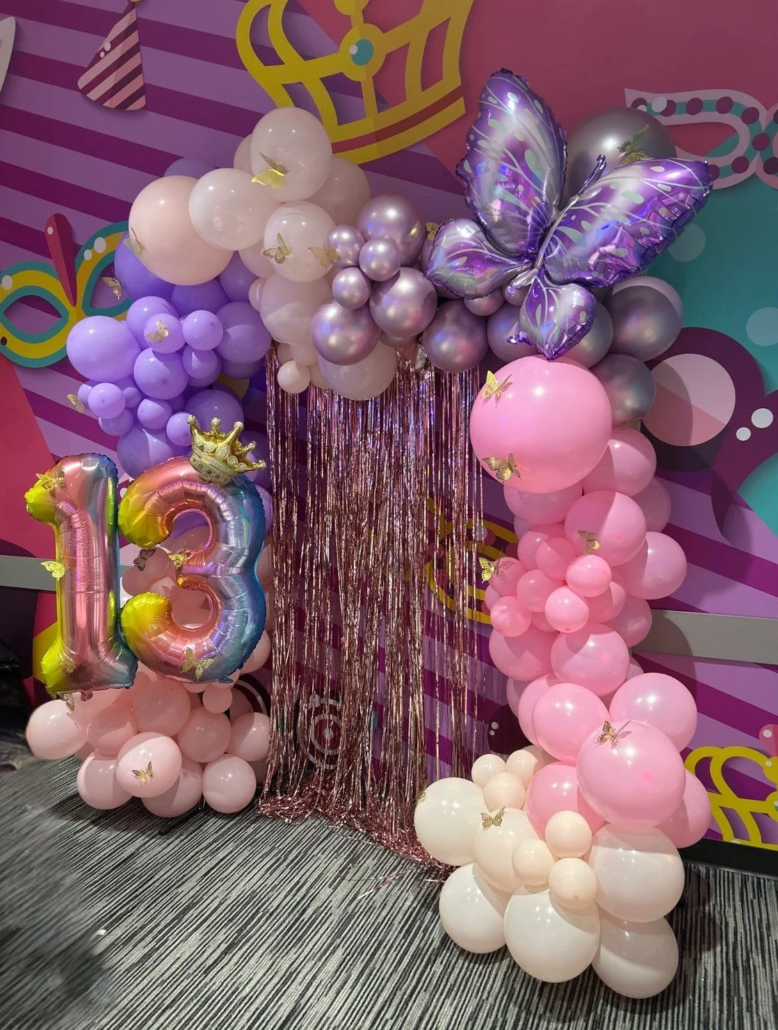 Balloon display with pink, purple, and white balloons, including butterfly and crown decorations, shaped as a rainbow and the number 13, with purple metallic butterfly balloon and pink shiny fringe backdrop, set against a colorful wall.