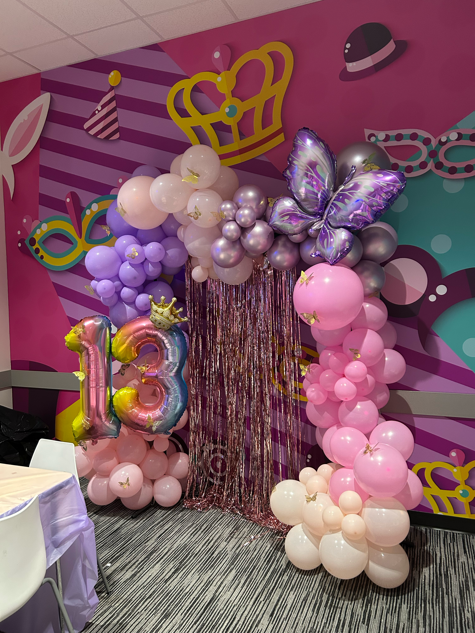 Pink, purple, and white balloons arranged with butterfly and crown decorations, with a shiny butterfly balloon and a large number 13 balloon, creating a festive birthday decoration backdrop in a colorful room.