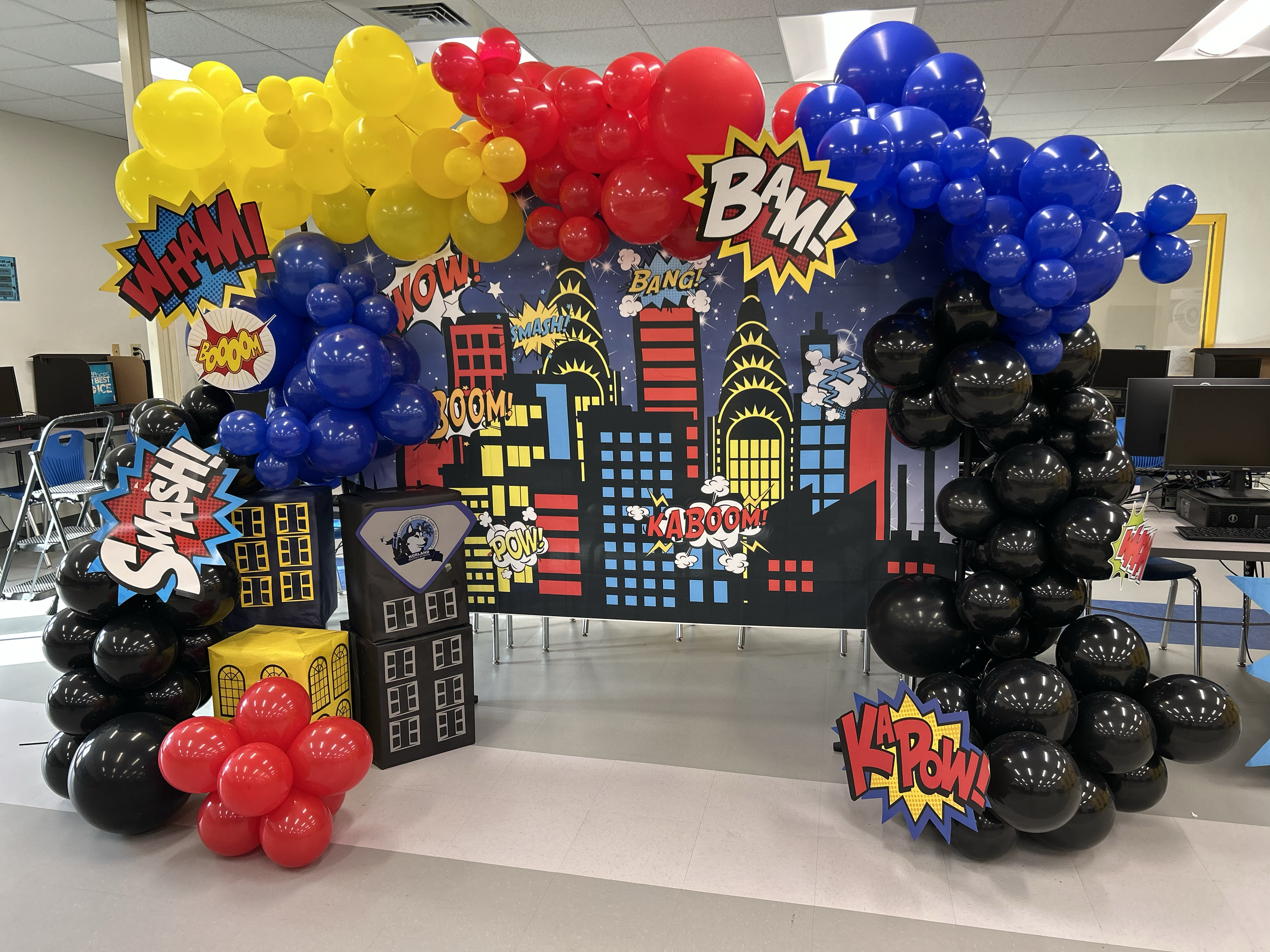 A superhero-themed birthday party photo booth with colorful balloons and comic book style cityscape backdrop.