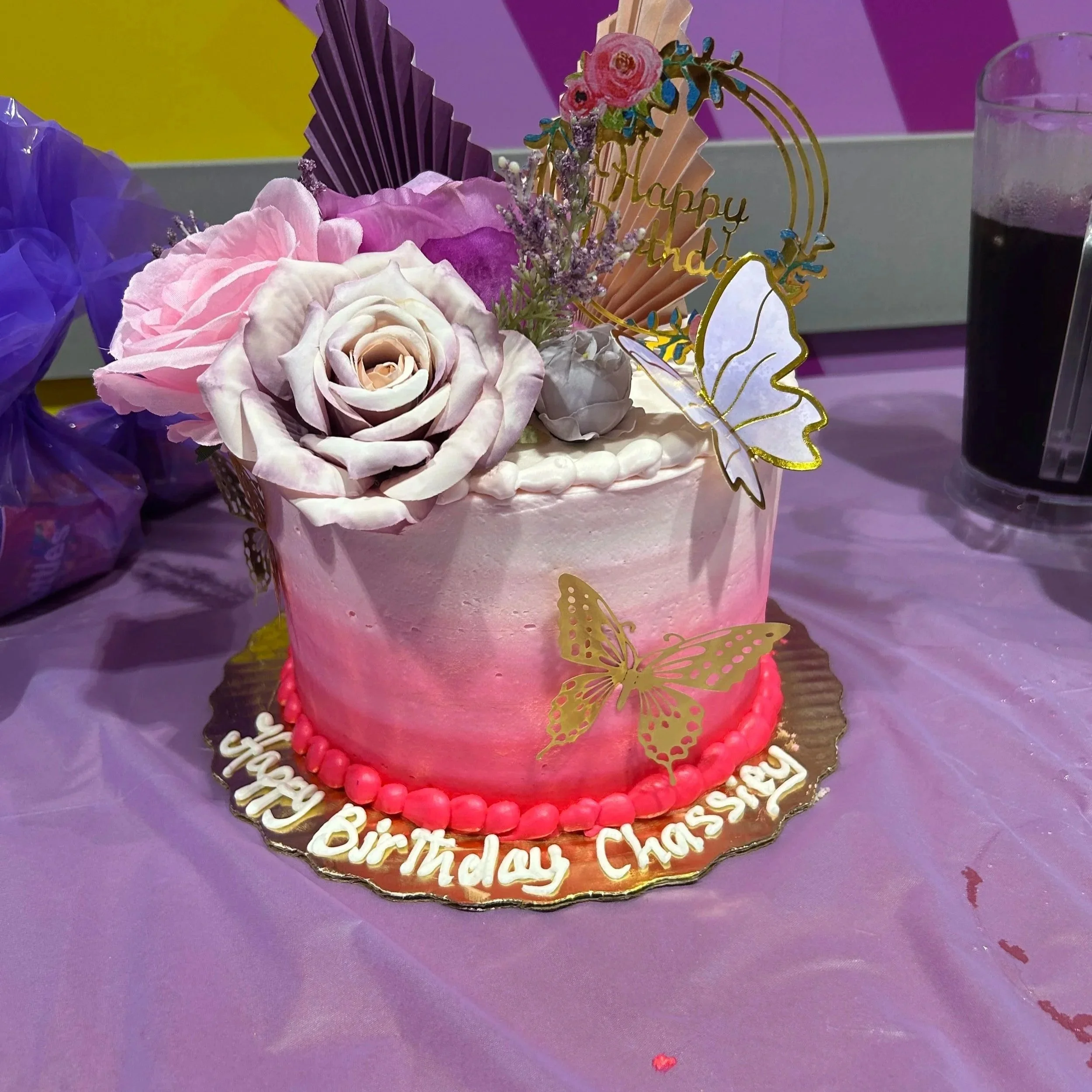 Pink birthday cake decorated with flowers, butterflies, and a "Happy Birthday" sign, placed on a purple tablecloth.