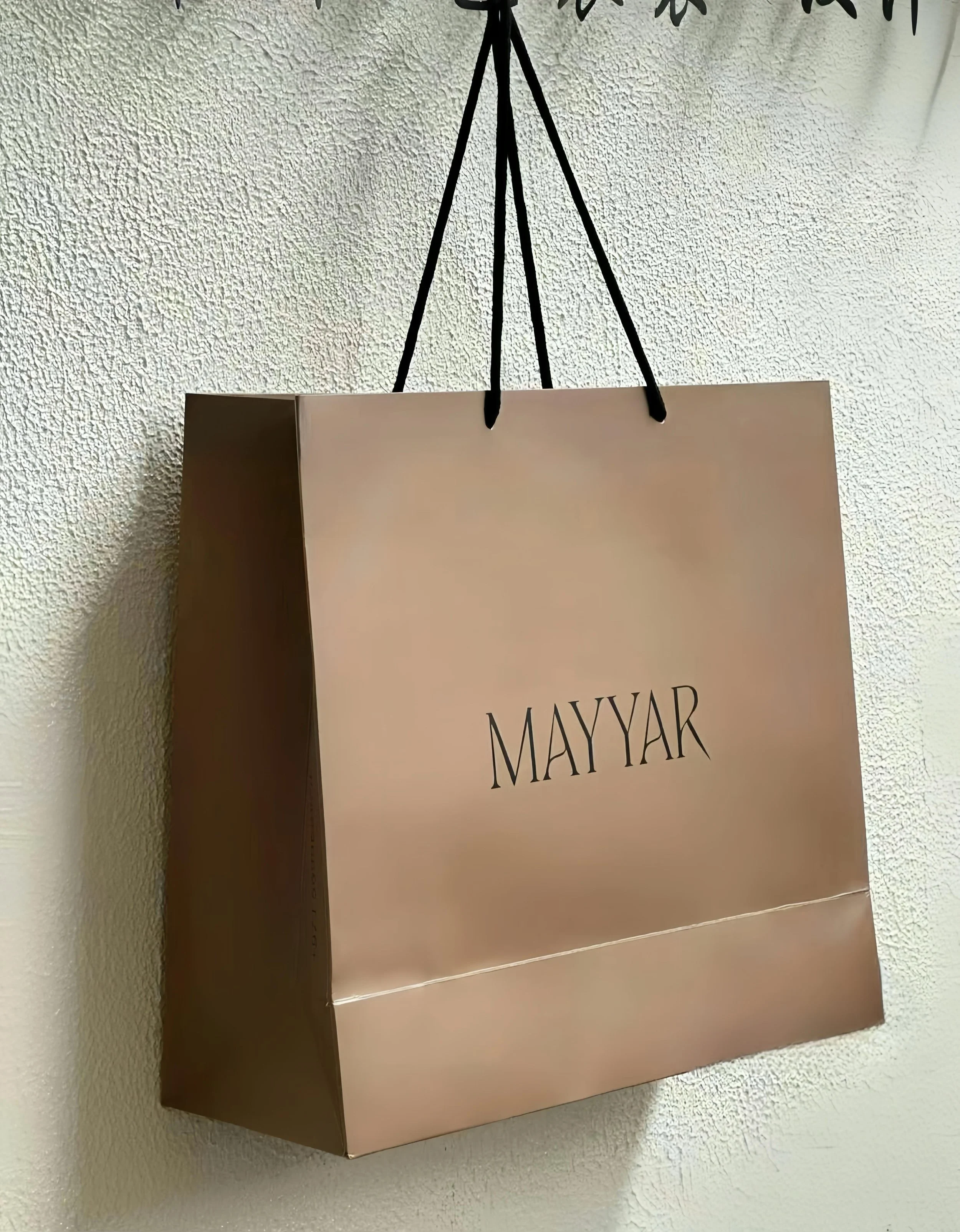 En rosa gåvokasse med texten MAYYAR, hängande i ett svart band mot en vit vägg.