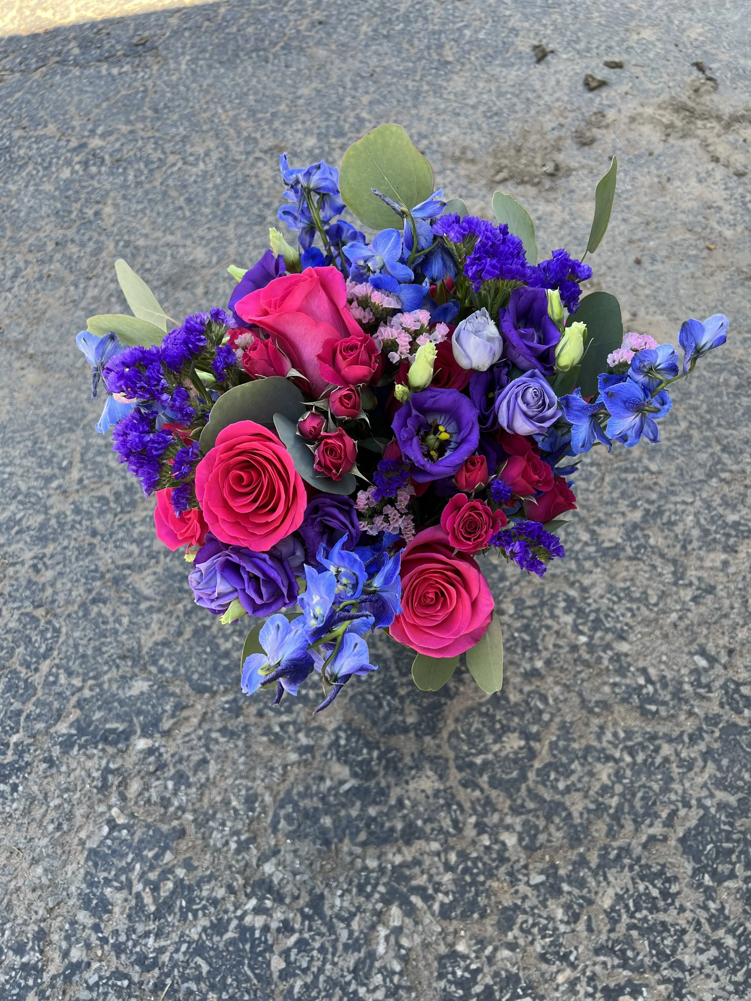 A colorful bouquet of flowers including pink roses, blue delphiniums, purple lisianthus, and green eucalyptus leaves, resting on a gravel ground.