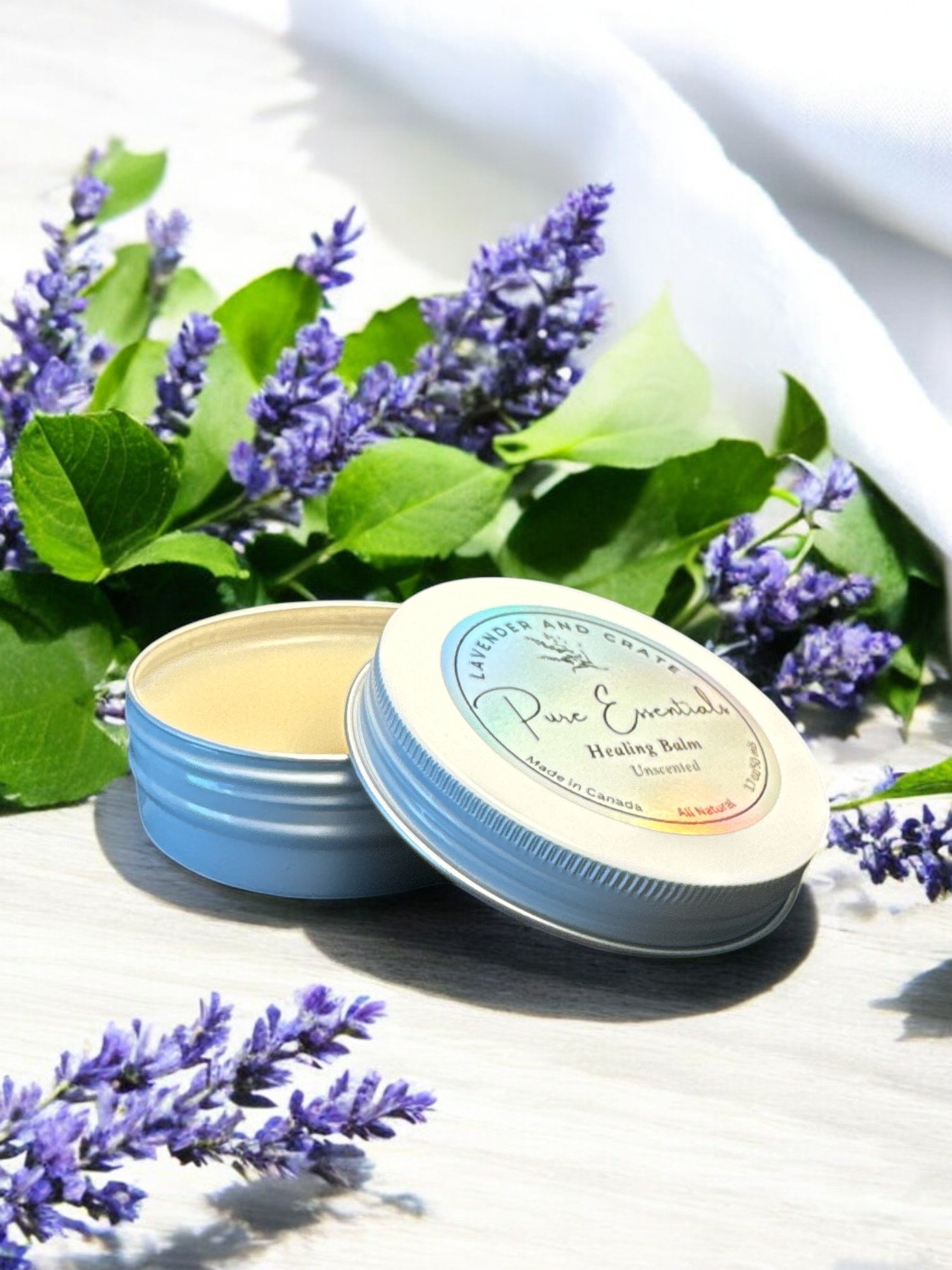 Pure Essentials Healing Balm in light blue tin with white label, surrounded by fresh purple lavender sprigs and green leaves on white surface, natural skincare product photography