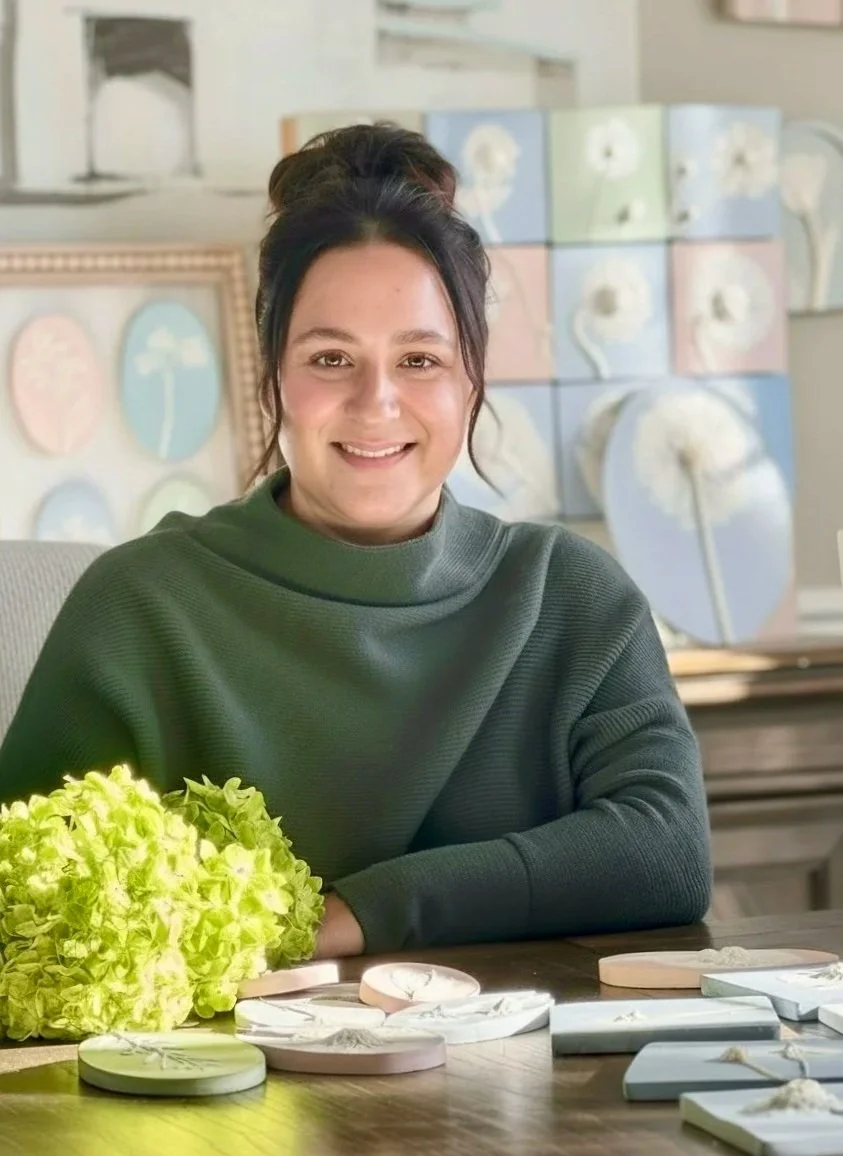 A woman with dark hair tied in a bun, wearing a green sweater, sitting at a table with a bouquet of green flowers and small decorative boxes. In the background, there are framed art pieces featuring dandelions and a wall with pastel-colored art.