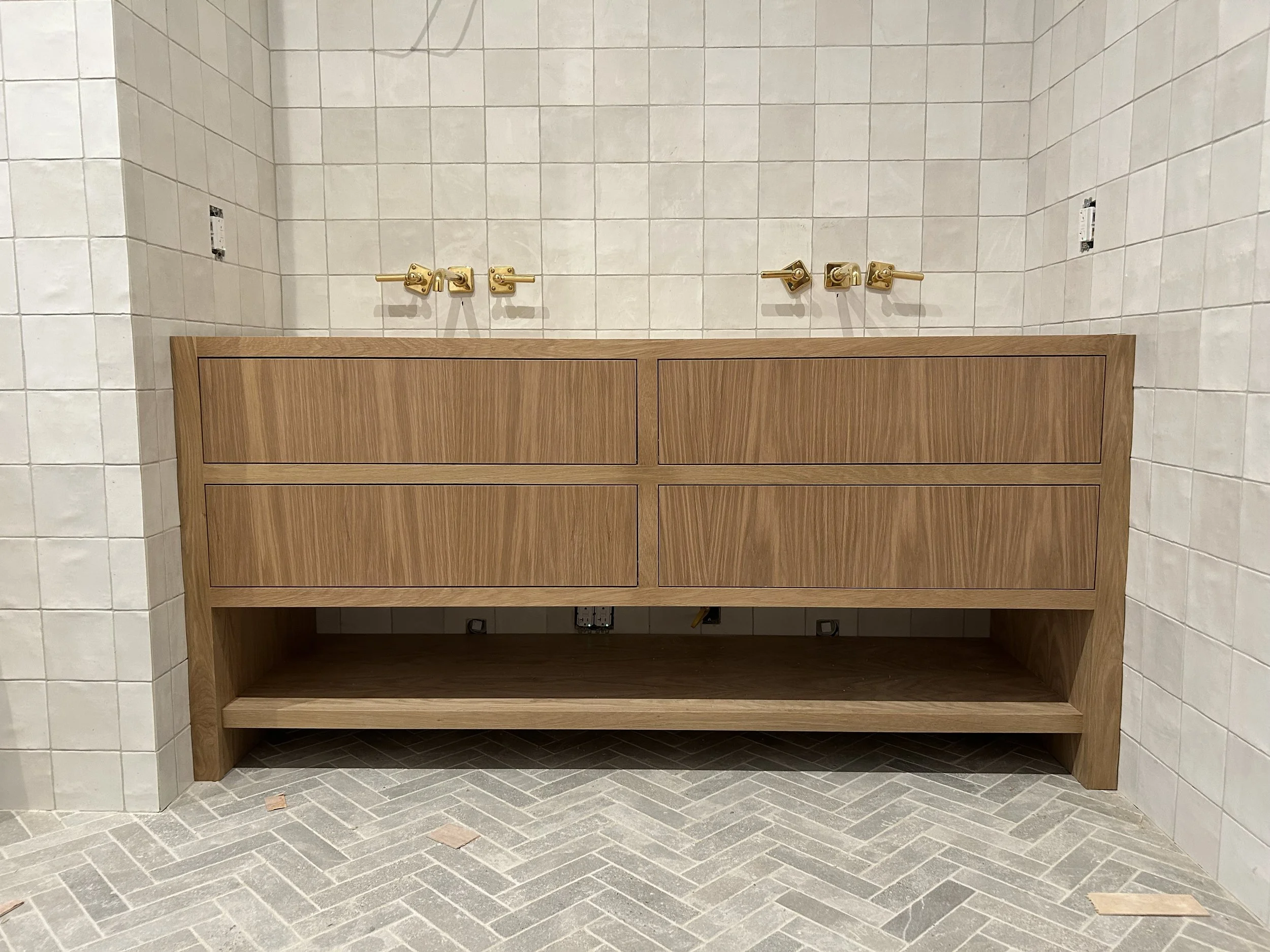 Wooden bathroom vanity with four drawers, a lower open shelf, and gold-colored faucet fixtures on a tiled wall, under construction or renovation.