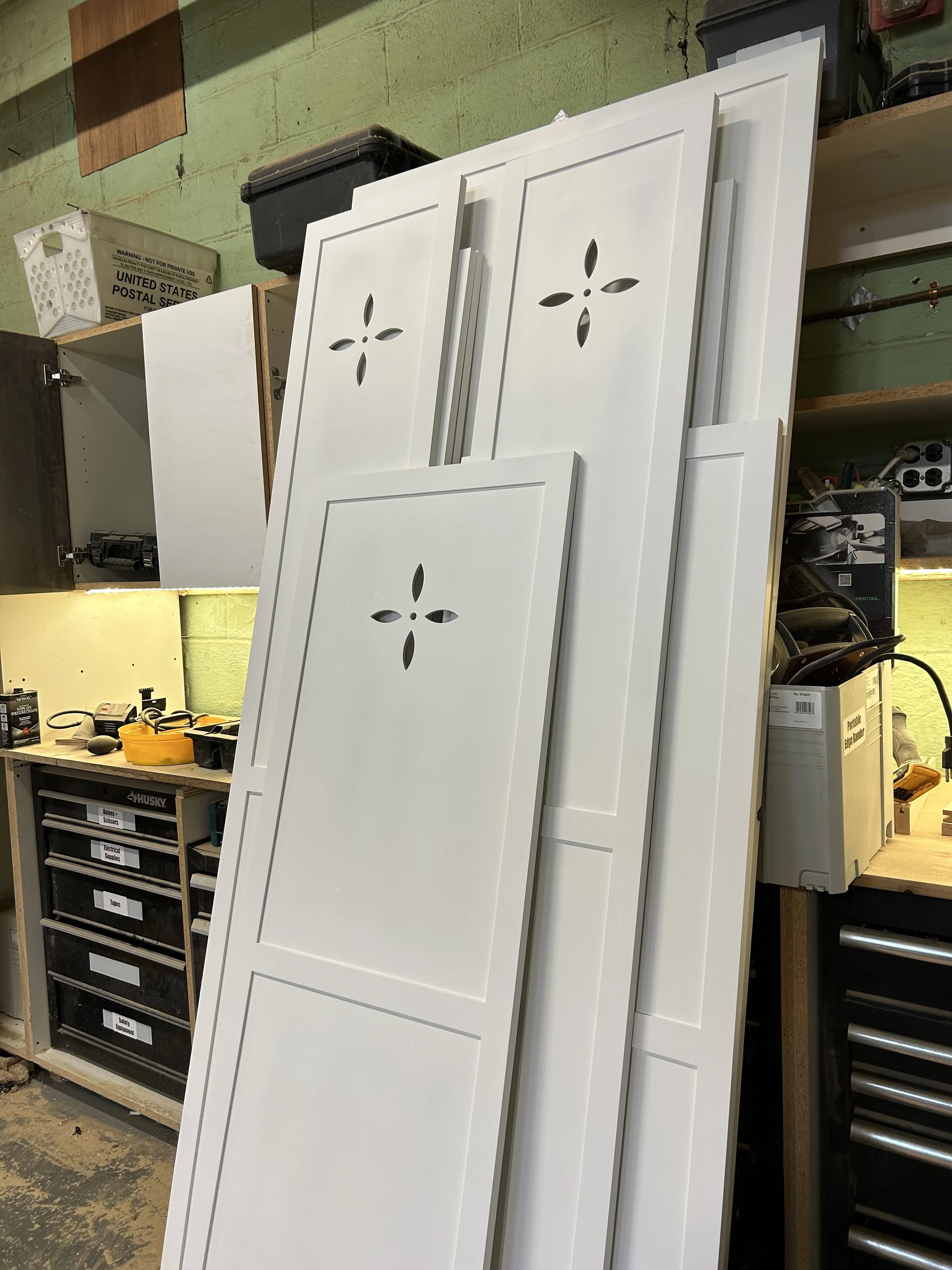 White cabinet doors with decorative cut-outs leaning against a workbench in a workshop.